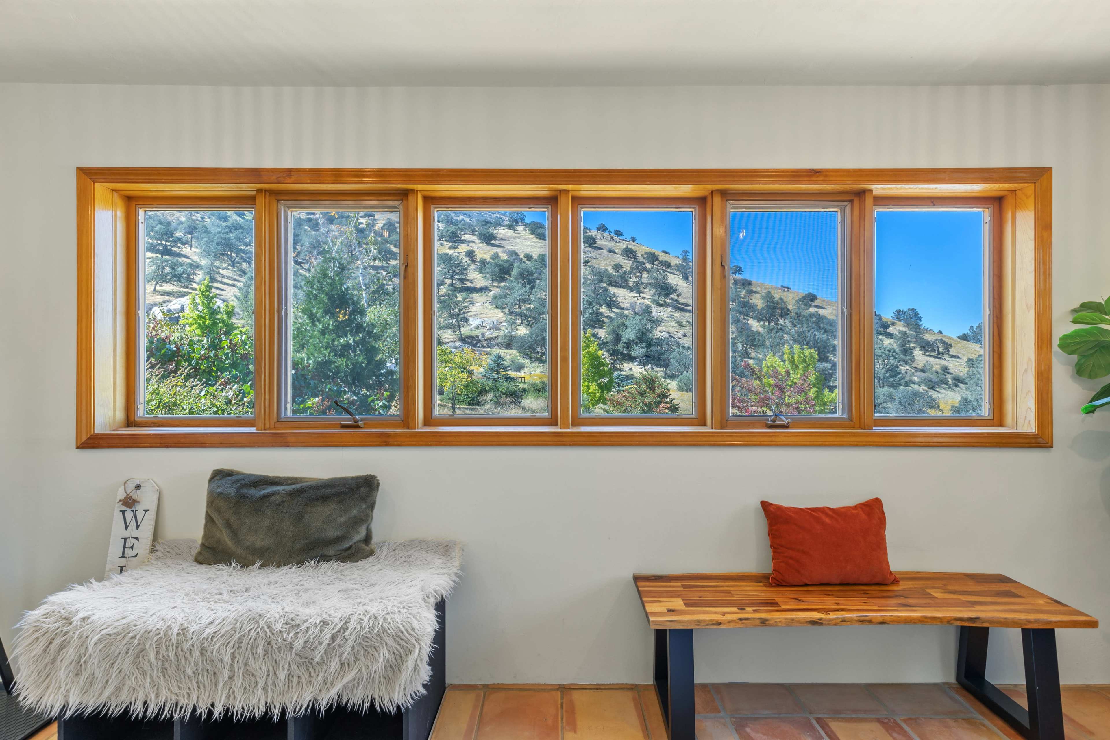 A cozy interior with a bench and a wooden table, framed by large windows that offer a view of a hillside.