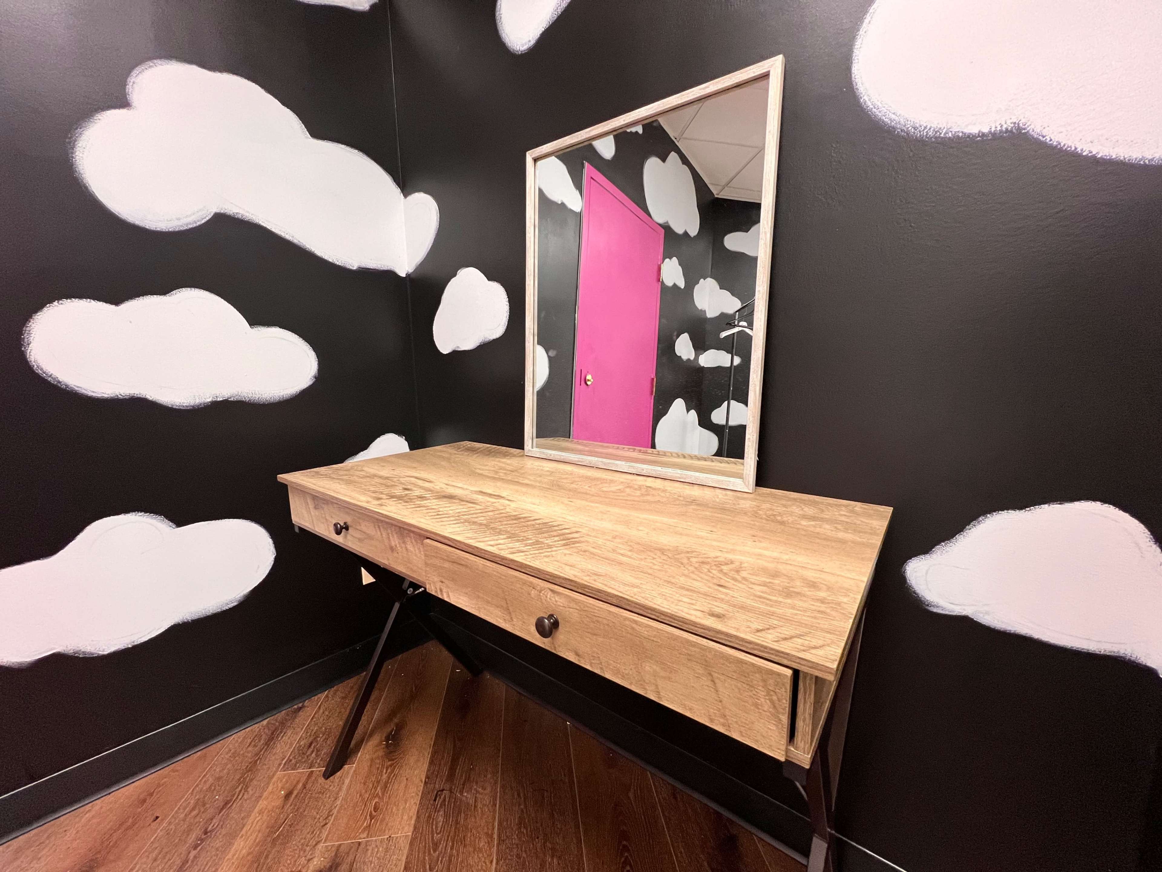 A wooden vanity table with a mirror is positioned against a black wall adorned with cloud-shaped designs and a pink door.