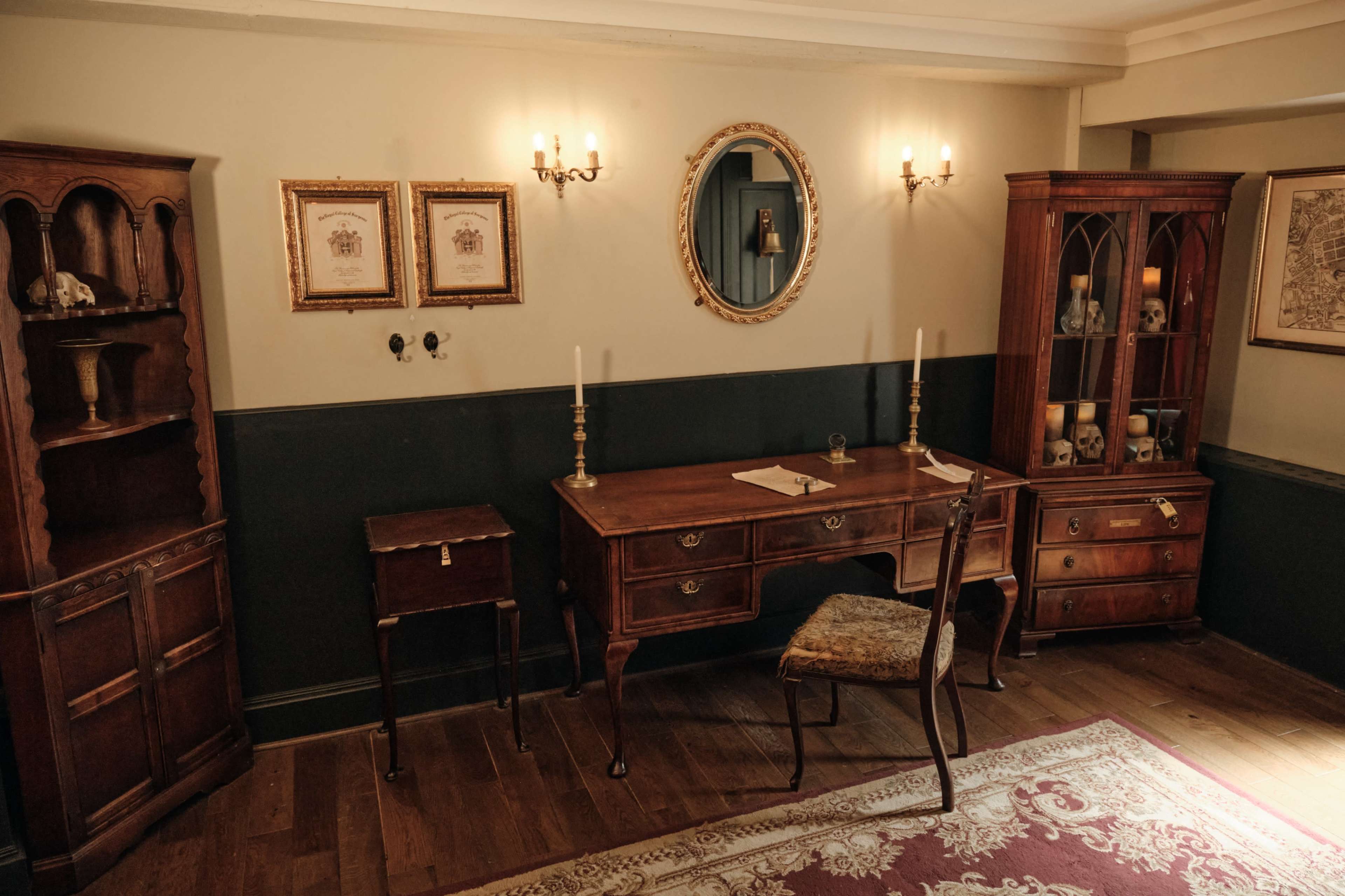 The image depicts a historical-style office with wooden furniture, including a desk and shelves, adorned with decorative items and framed pictures on the walls.
