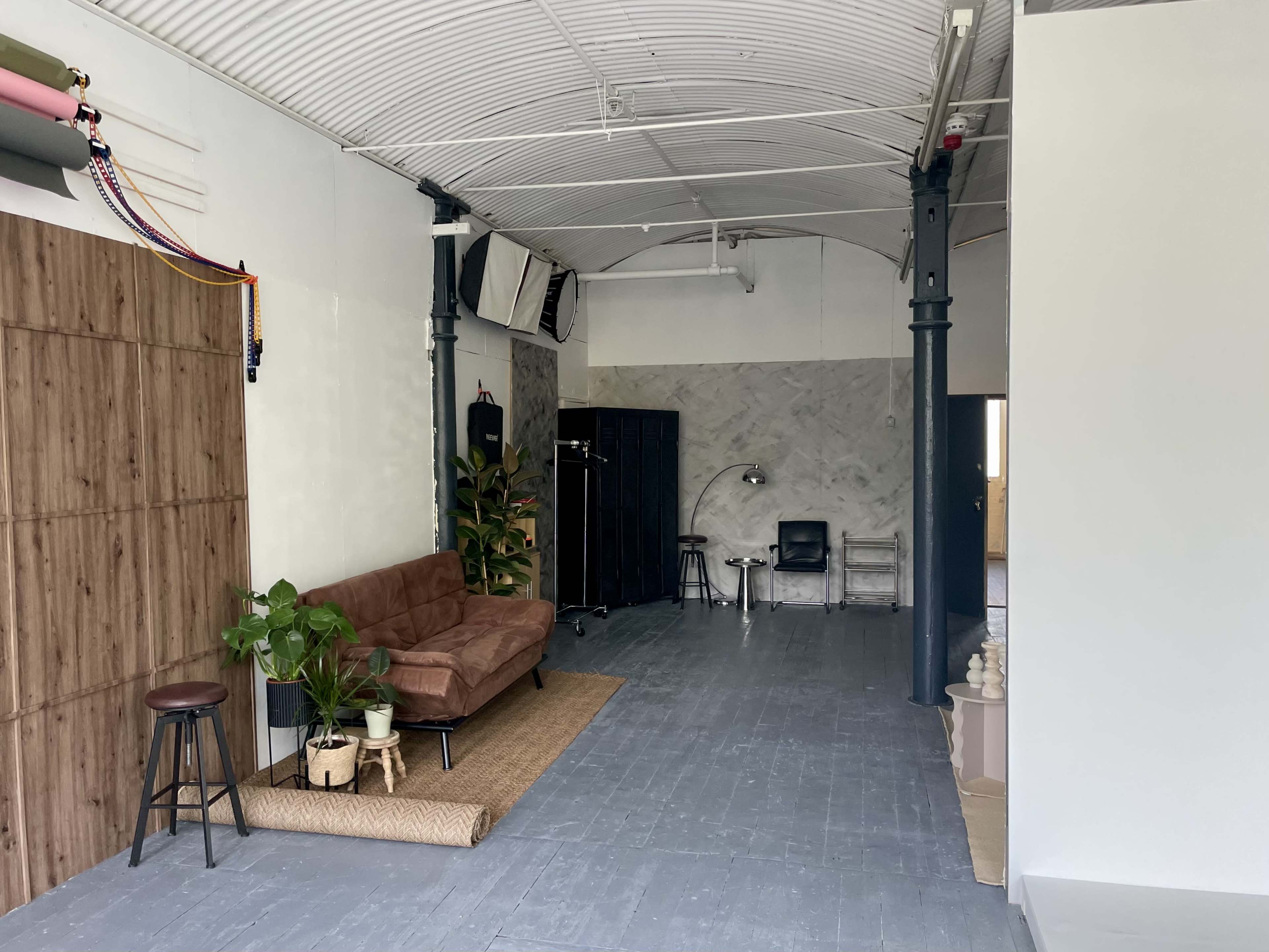 The image shows a spacious interior with a brown couch, a plant, a stool, and various furnishings against a light-colored wall.