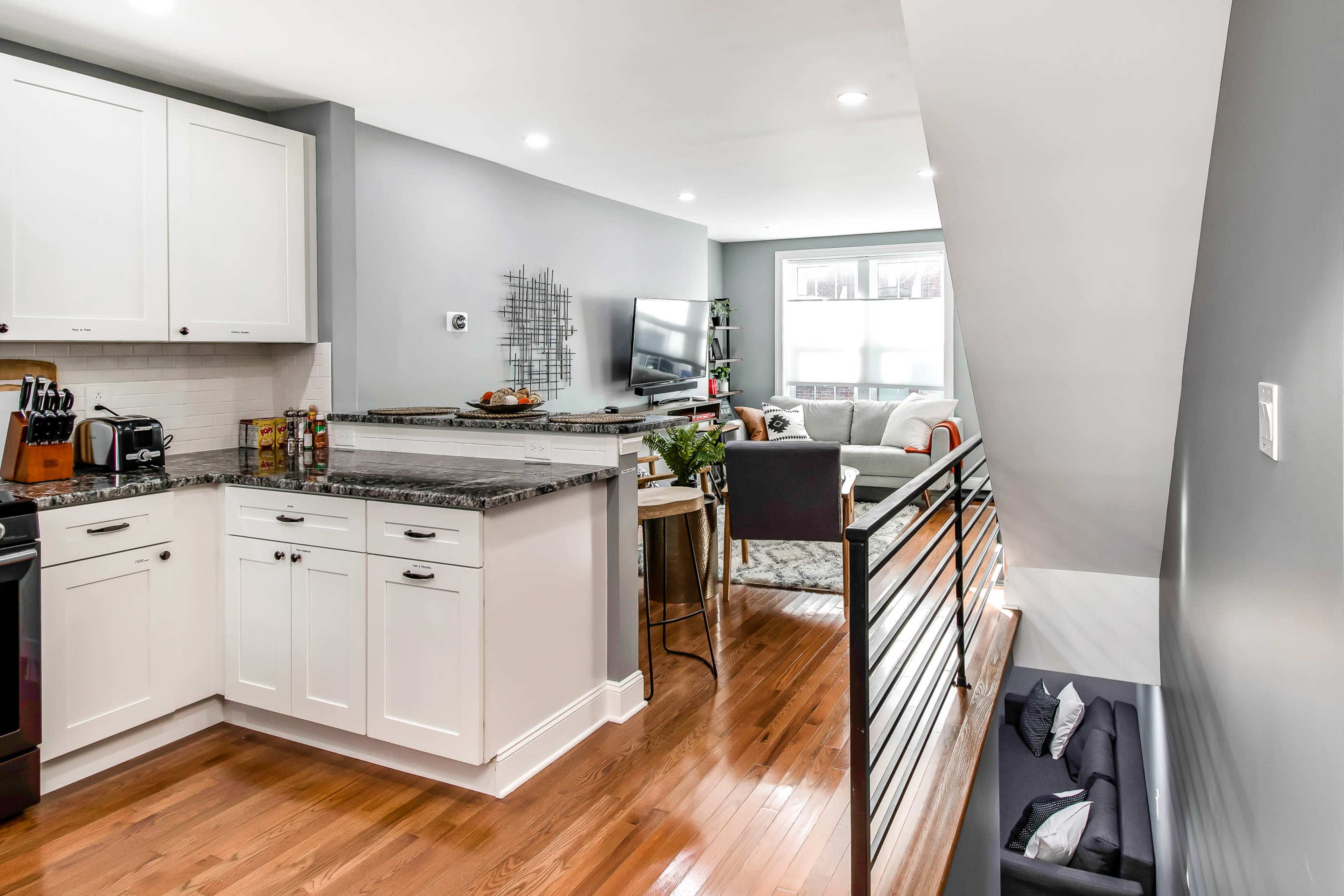 A modern kitchen with white cabinets and granite countertops is seamlessly connected to a living area featuring a sofa and television, all within a well-lit space.