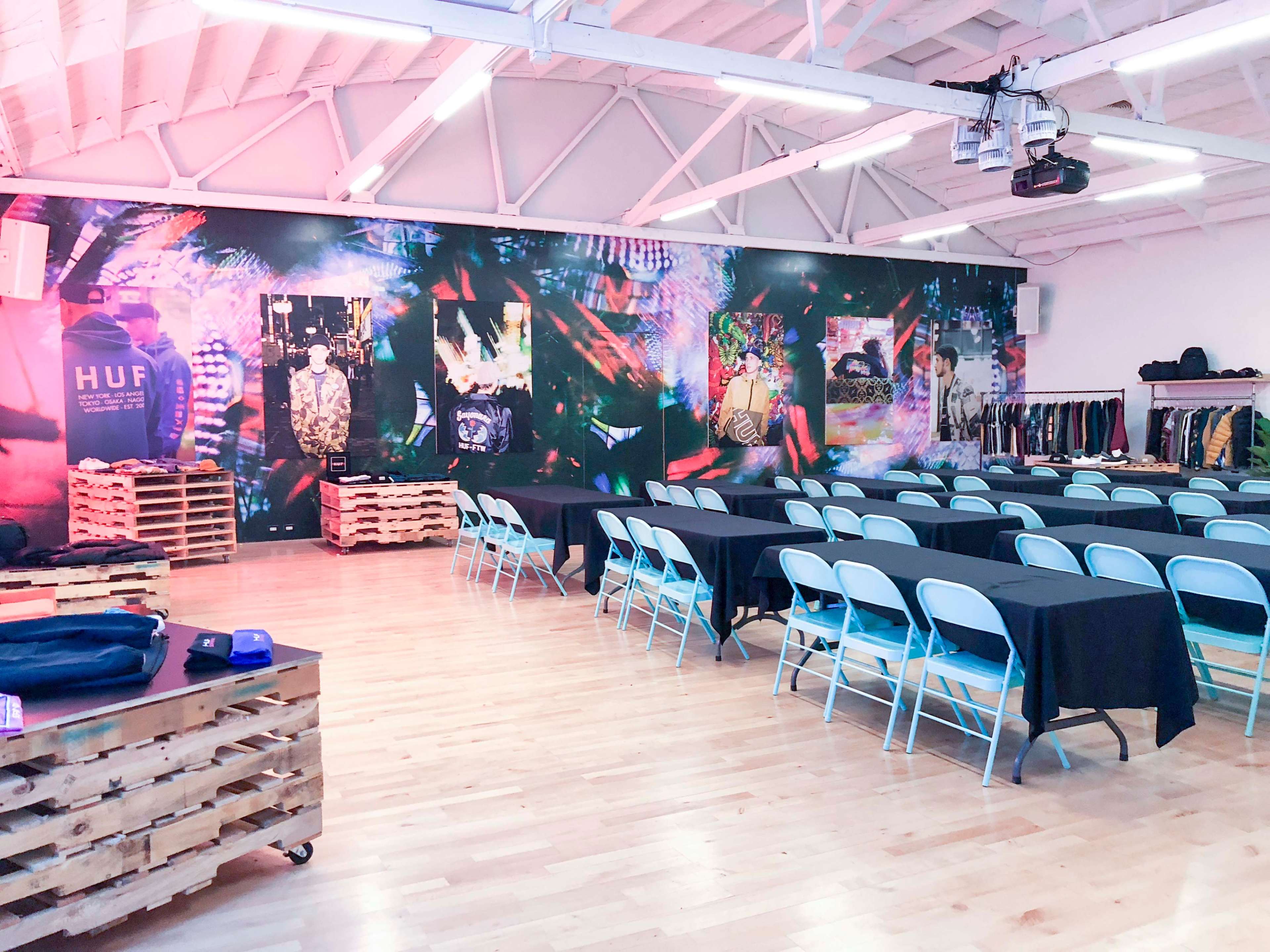 A spacious room features multiple tables covered with black cloth, surrounded by blue chairs, against a vividly decorated wall adorned with colorful graphics and images.