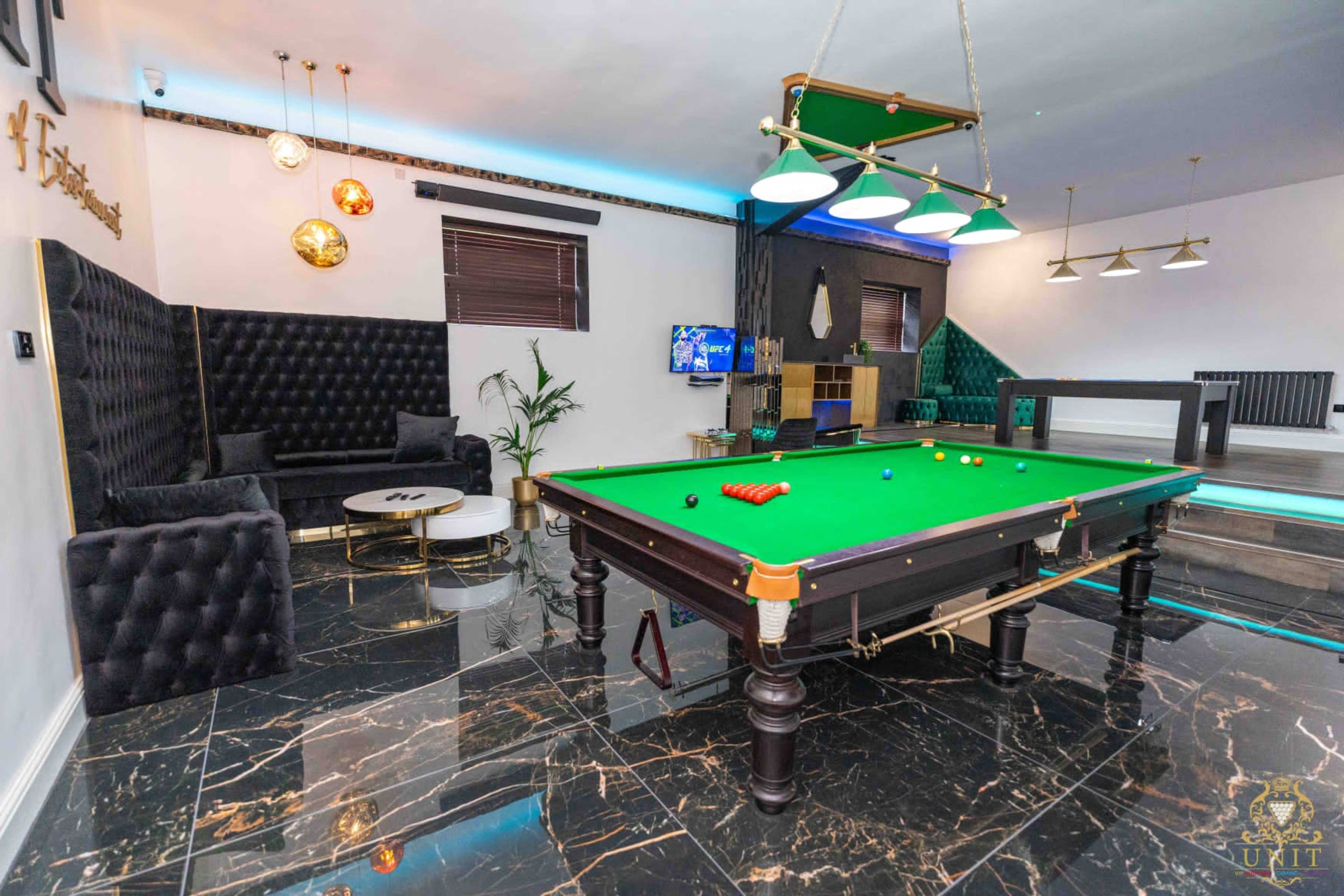 A game room features a green pool table surrounded by black and white furniture, with modern lighting and marble flooring.