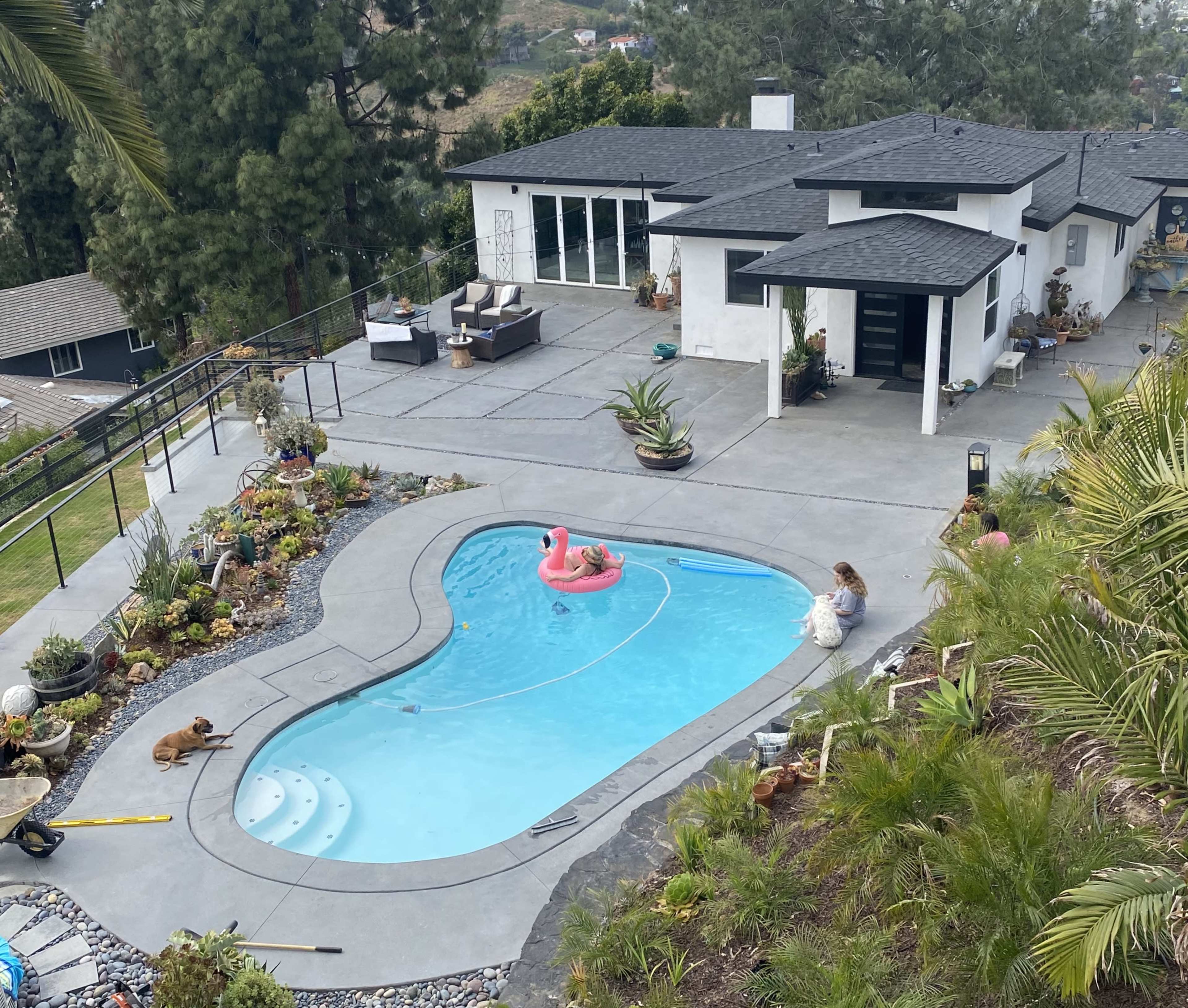 A person sits by a swimming pool shaped like a large kidney, while another swims with a pink float, surrounded by a landscaped yard and a modern house.