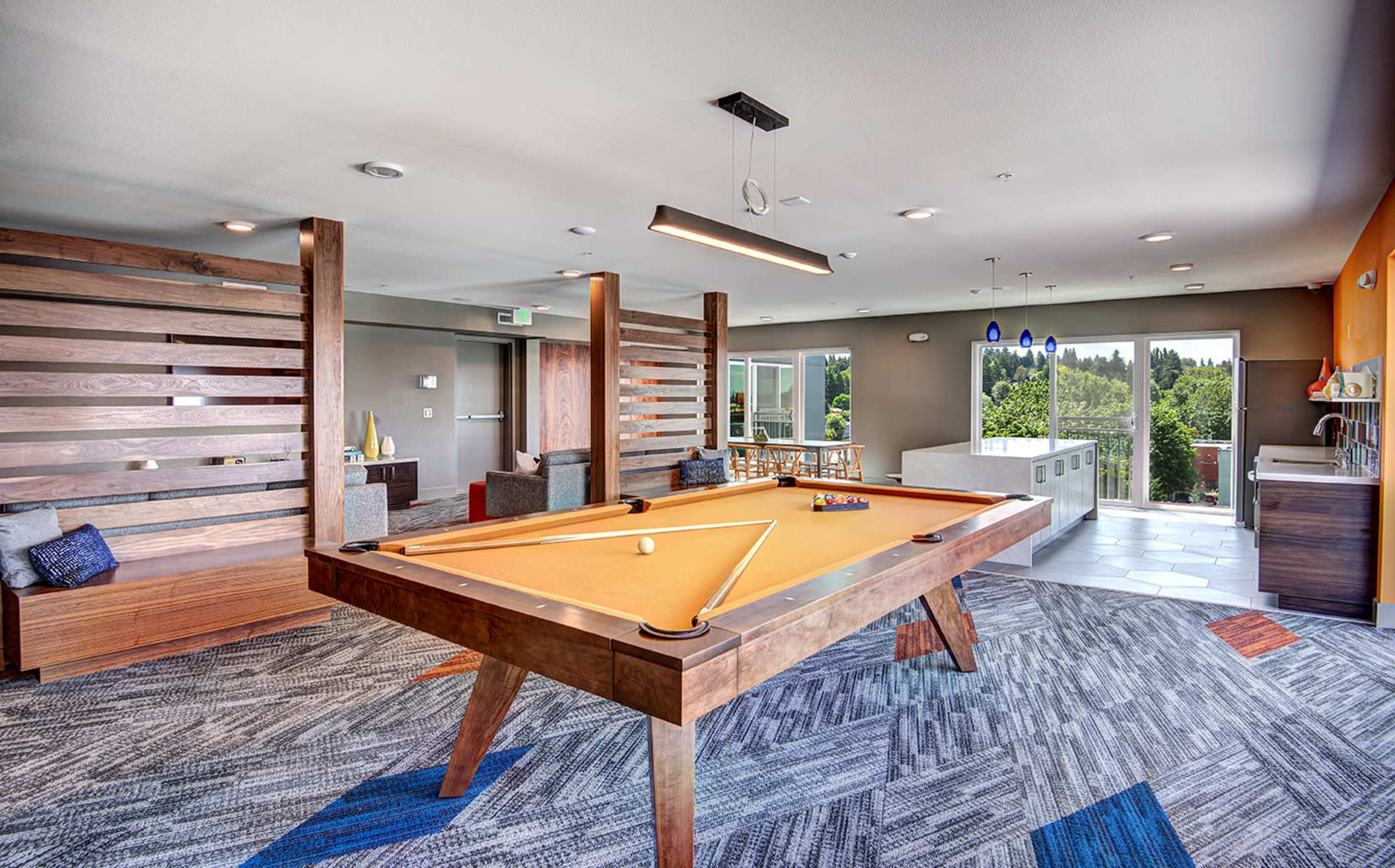 A modern lounge area features a wooden pool table surrounded by seating and large windows that provide natural light and a view of the outdoors.