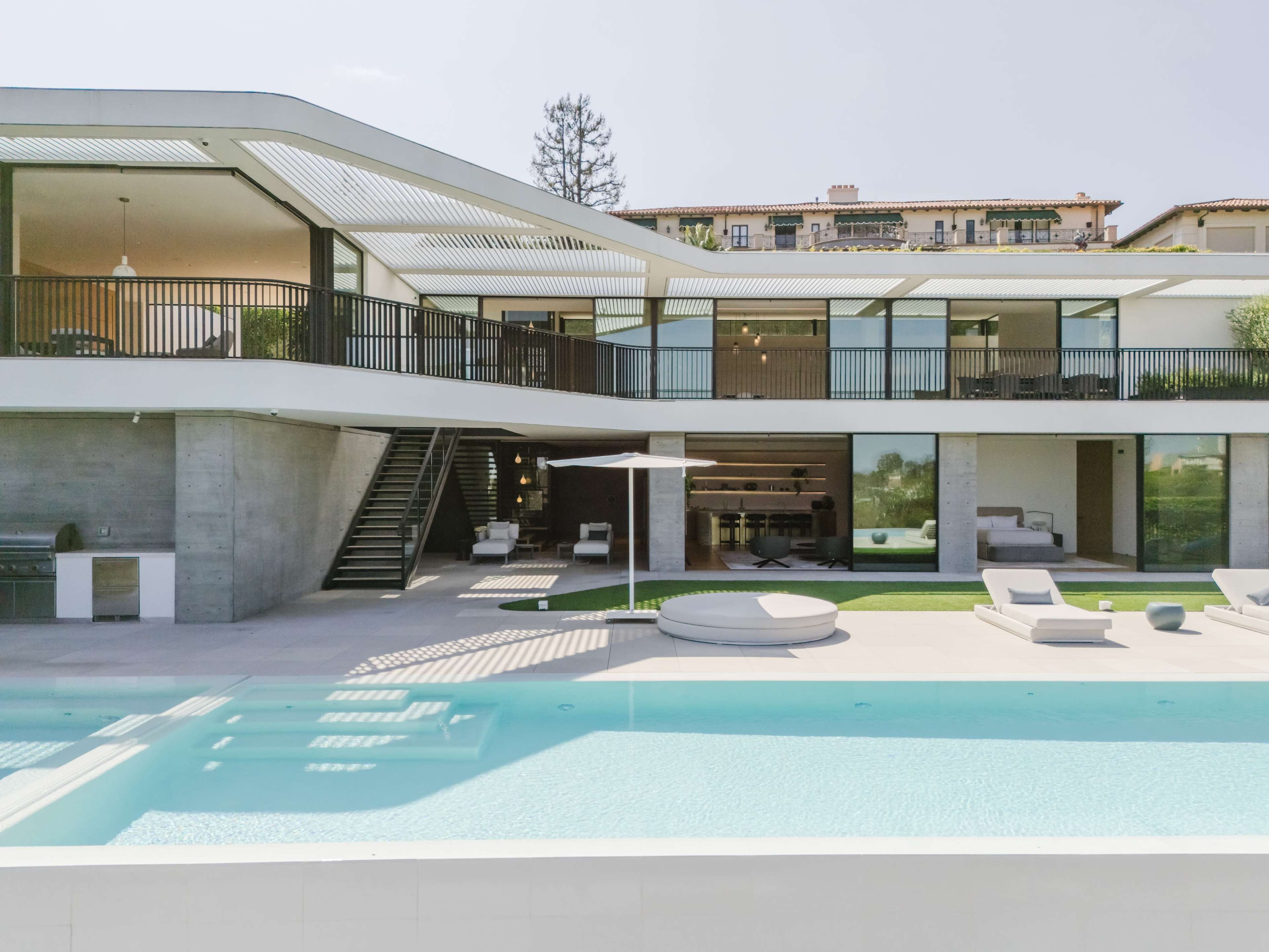 A modern two-story house features a large swimming pool, outdoor seating areas, and expansive windows that overlook a landscaped yard.