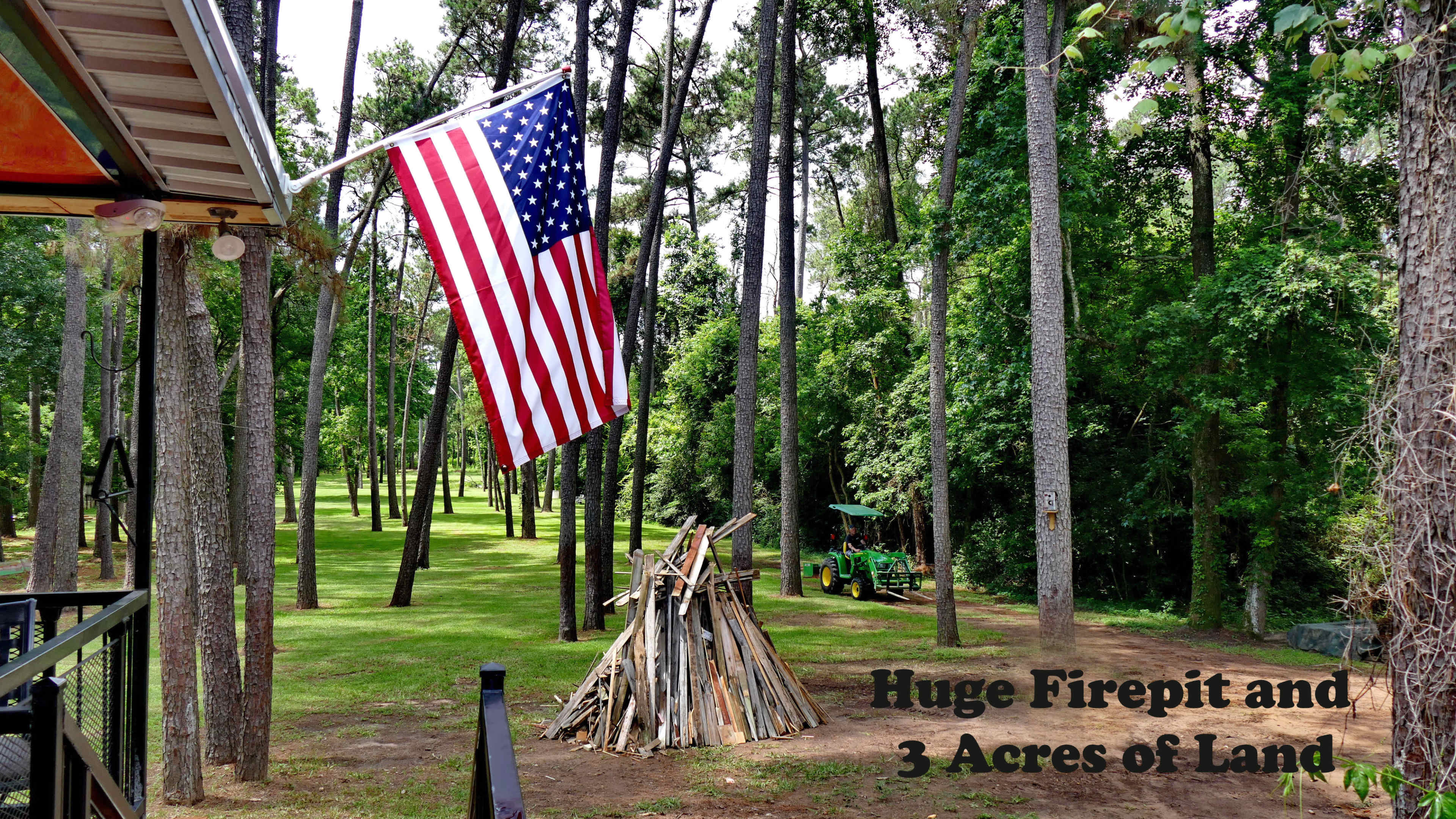 An American flag waving beside a large firepit and a pile of wood, set within a grassy area surrounded by trees on a three-acre property.