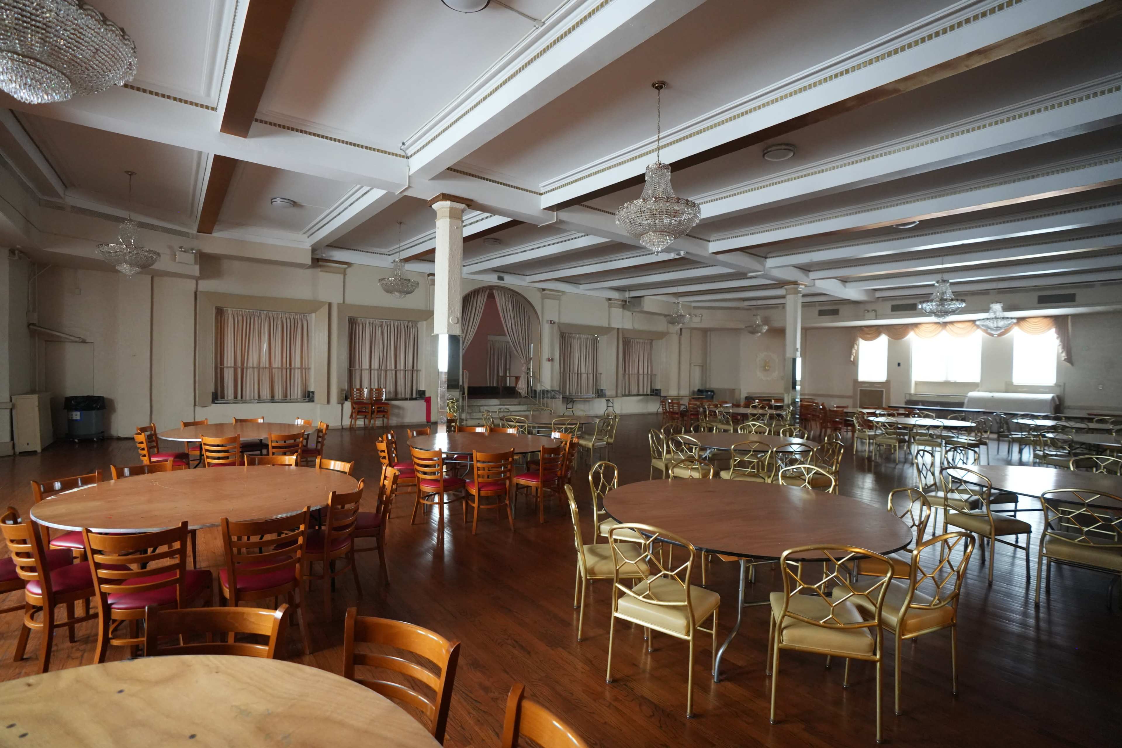 The image shows a spacious, empty hall arranged with several round dining tables and chairs, featuring chandeliers overhead.