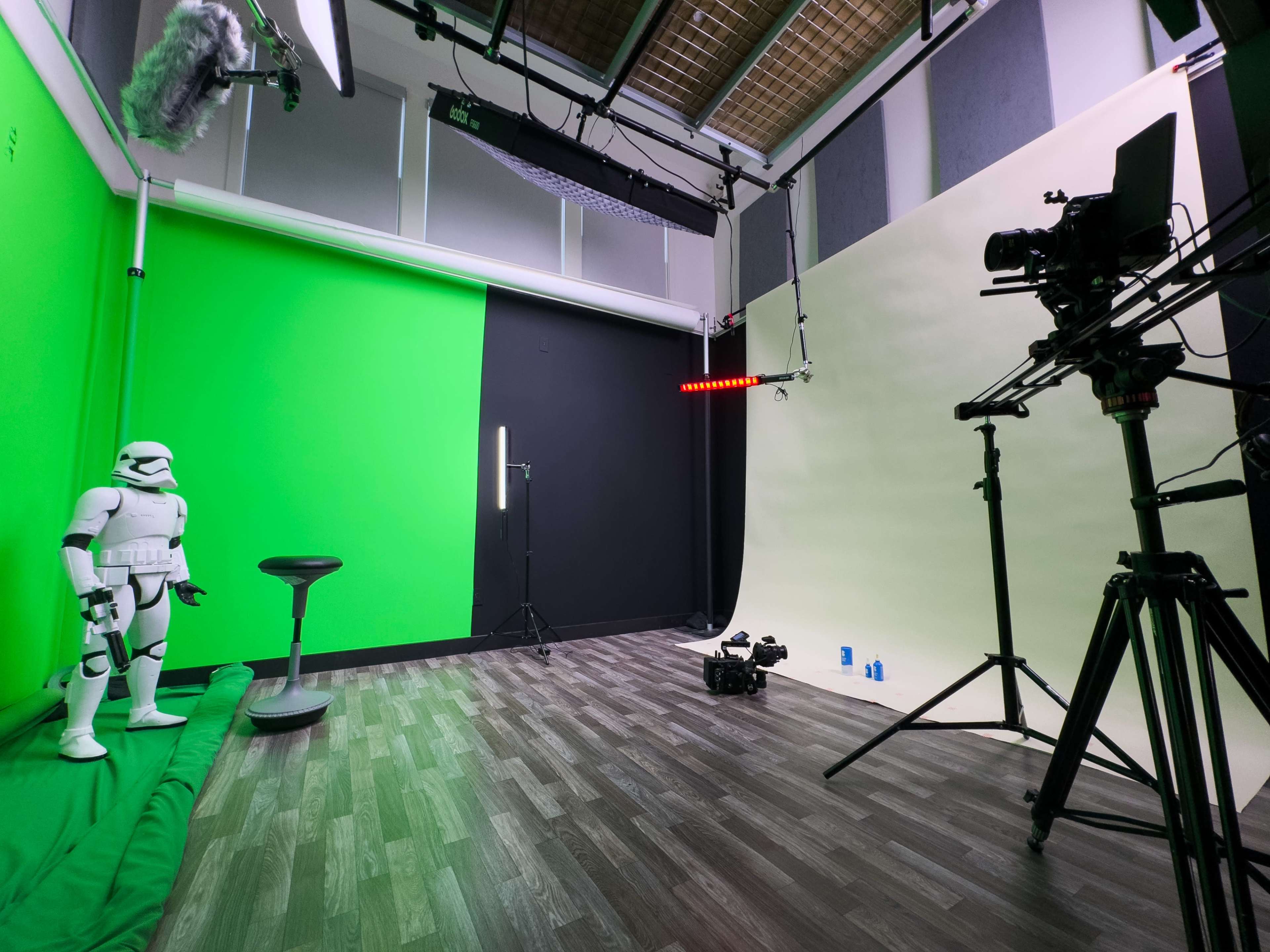 A film production studio featuring a green screen, a black wall, a beige backdrop, a camera on a tripod, lighting equipment, and a stormtrooper figure standing beside a stool.