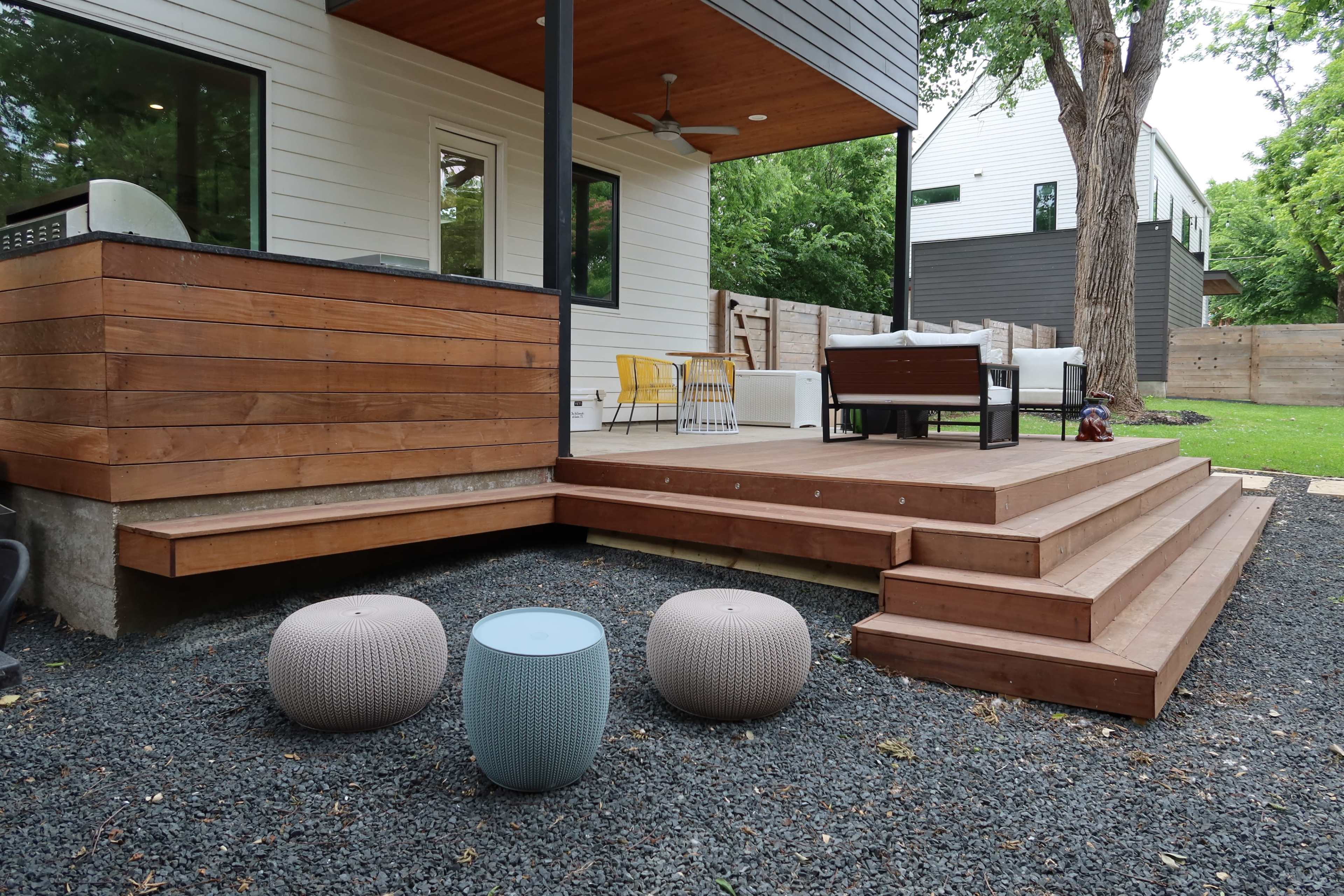 A wooden deck with steps leading down to a gravel area, featuring three round stools and a seating arrangement in a modern outdoor space.