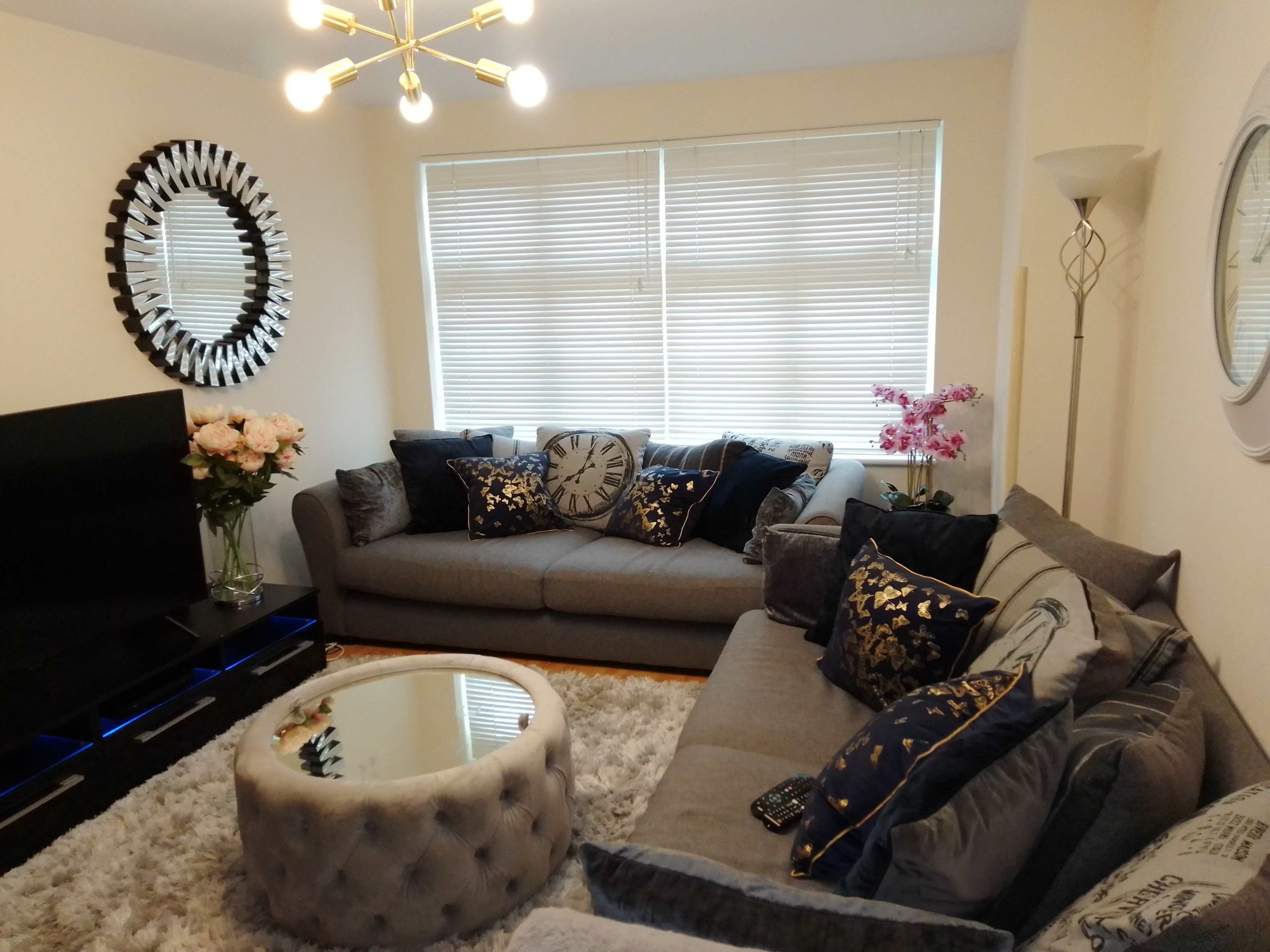 A modern living room featuring a gray sectional sofa, a round coffee table, a large mirror on the wall, and decorative pillows, with a floral arrangement nearby.