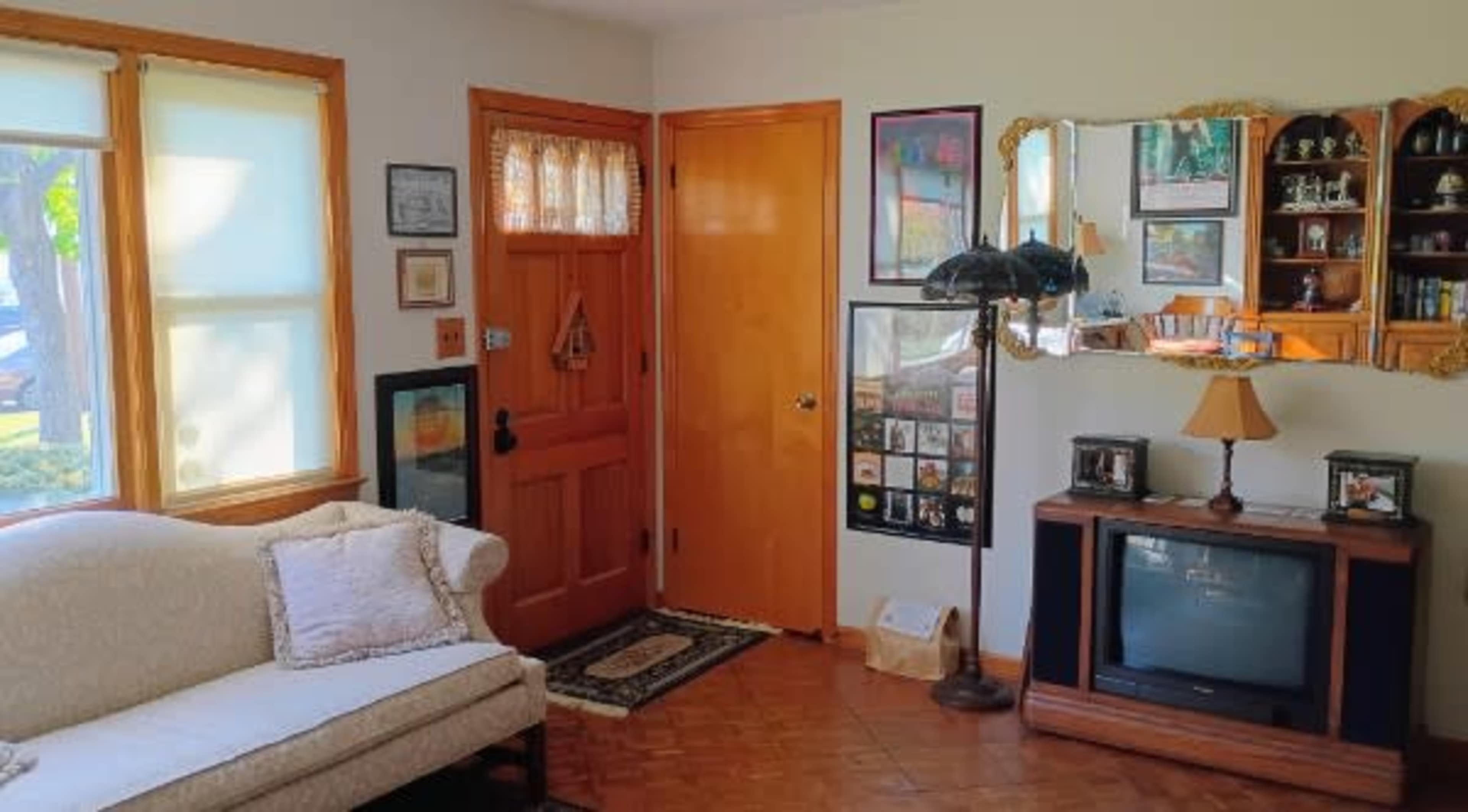 The image shows a cozy living room featuring a beige sofa, a wooden door with a decorative panel, and a wall adorned with framed pictures and a television set.