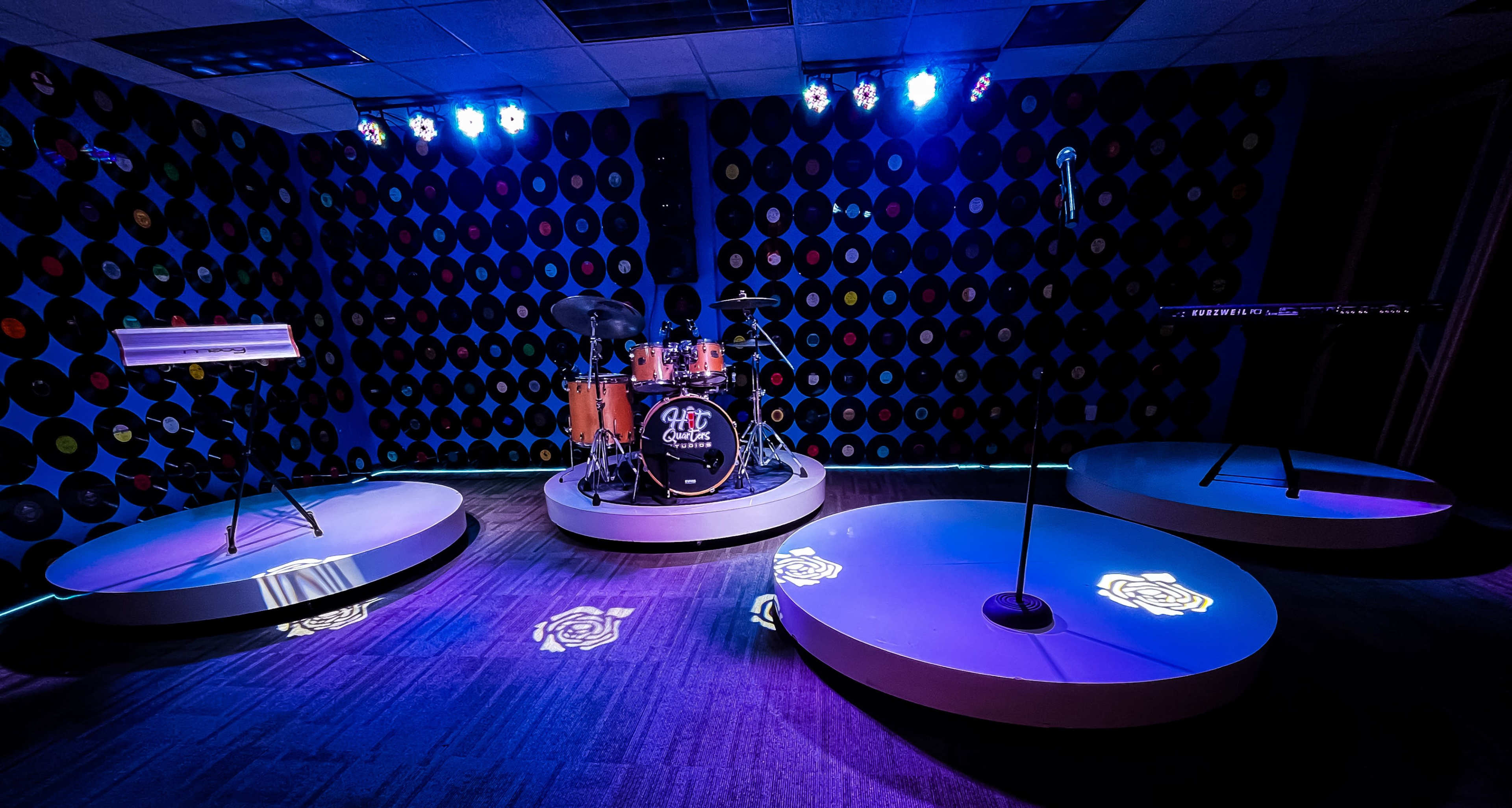 A music rehearsal space featuring a drum set, keyboard, and microphone stands arranged on circular platforms, illuminated by blue stage lighting against a backdrop of vinyl records.