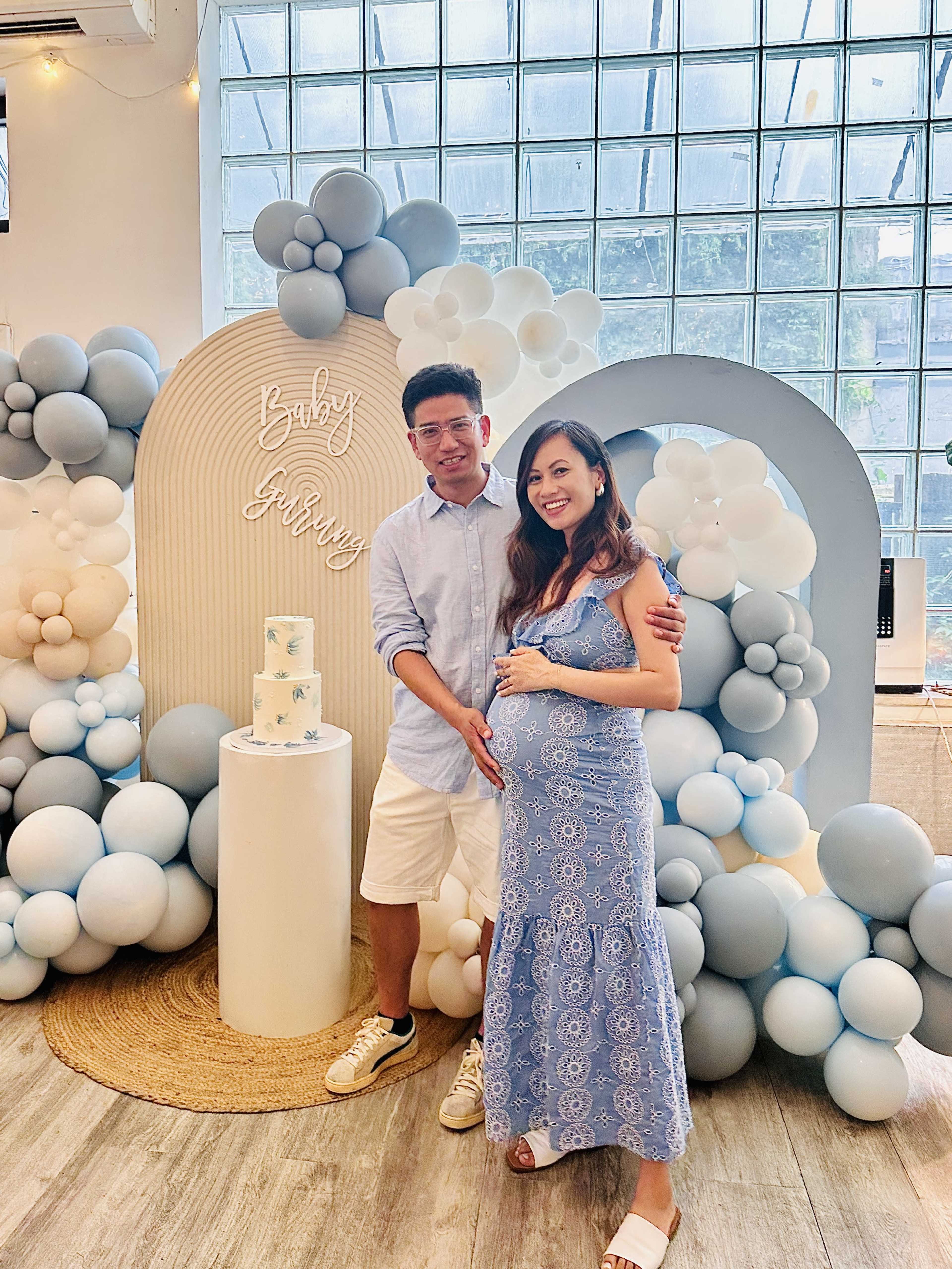 A couple stands next to a decorative display featuring balloons and a cake, celebrating a baby shower.