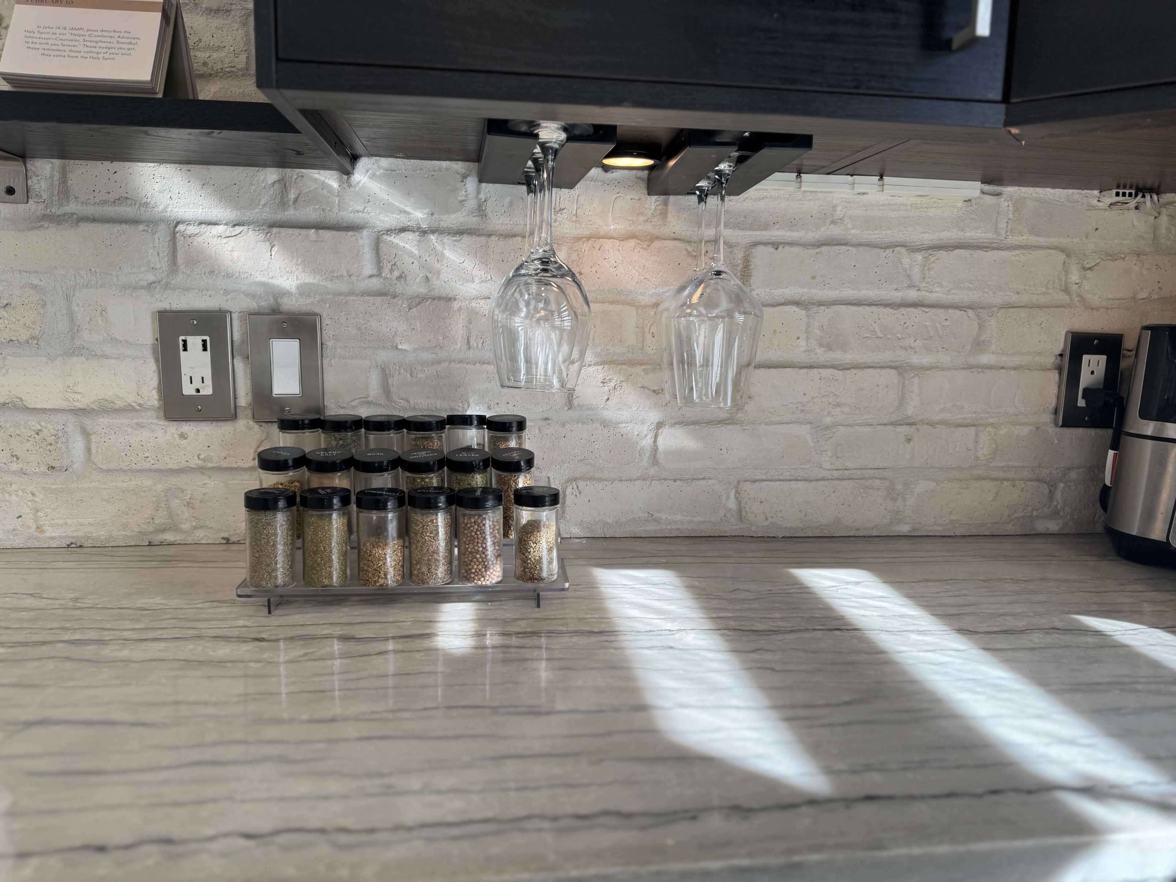 The image shows a kitchen countertop with a spice rack containing several jars and hanging wine glasses above it.