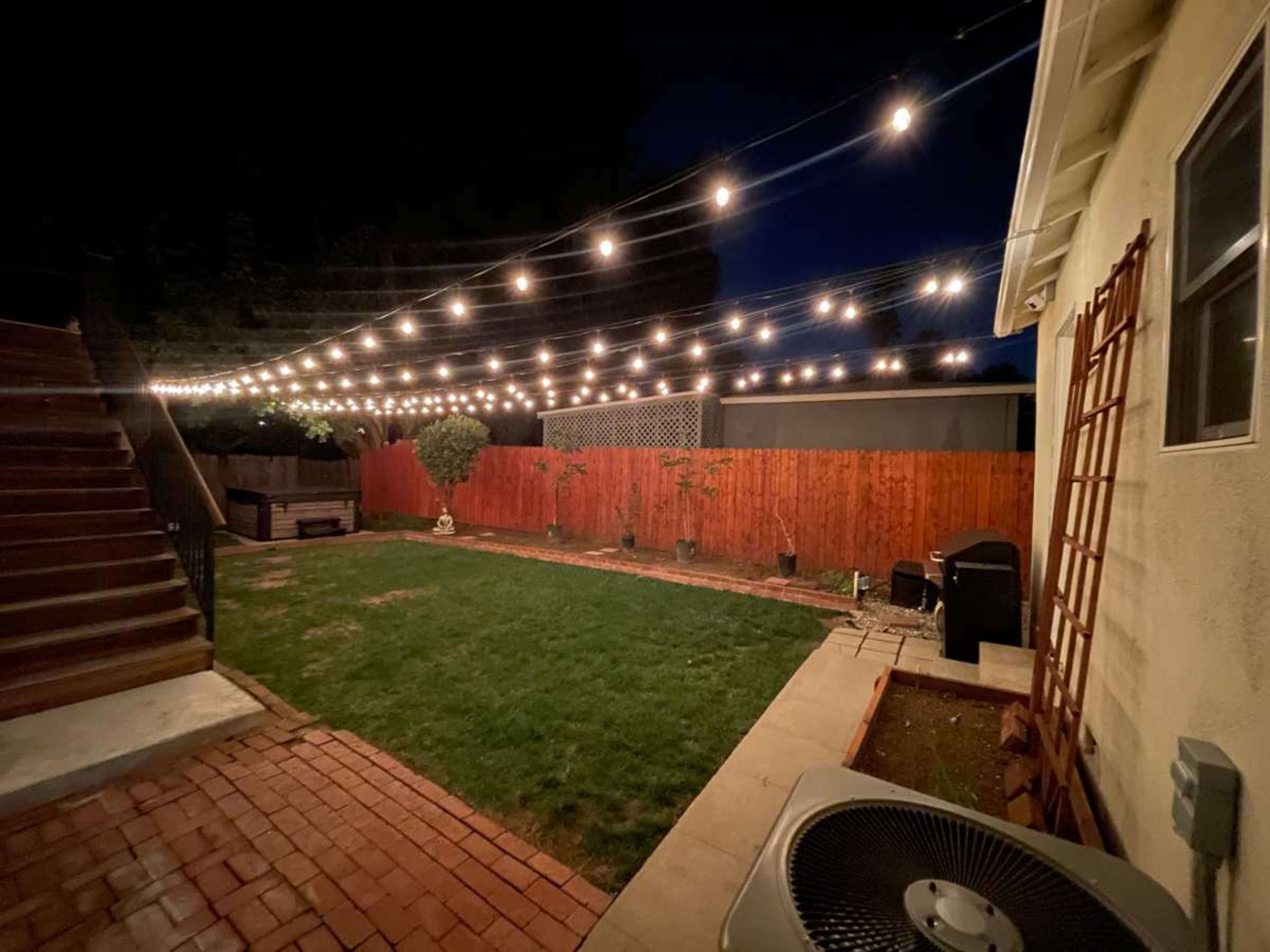 A backyard at night features string lights overhead, a grassy area, brick and concrete paths, and wooden fencing.