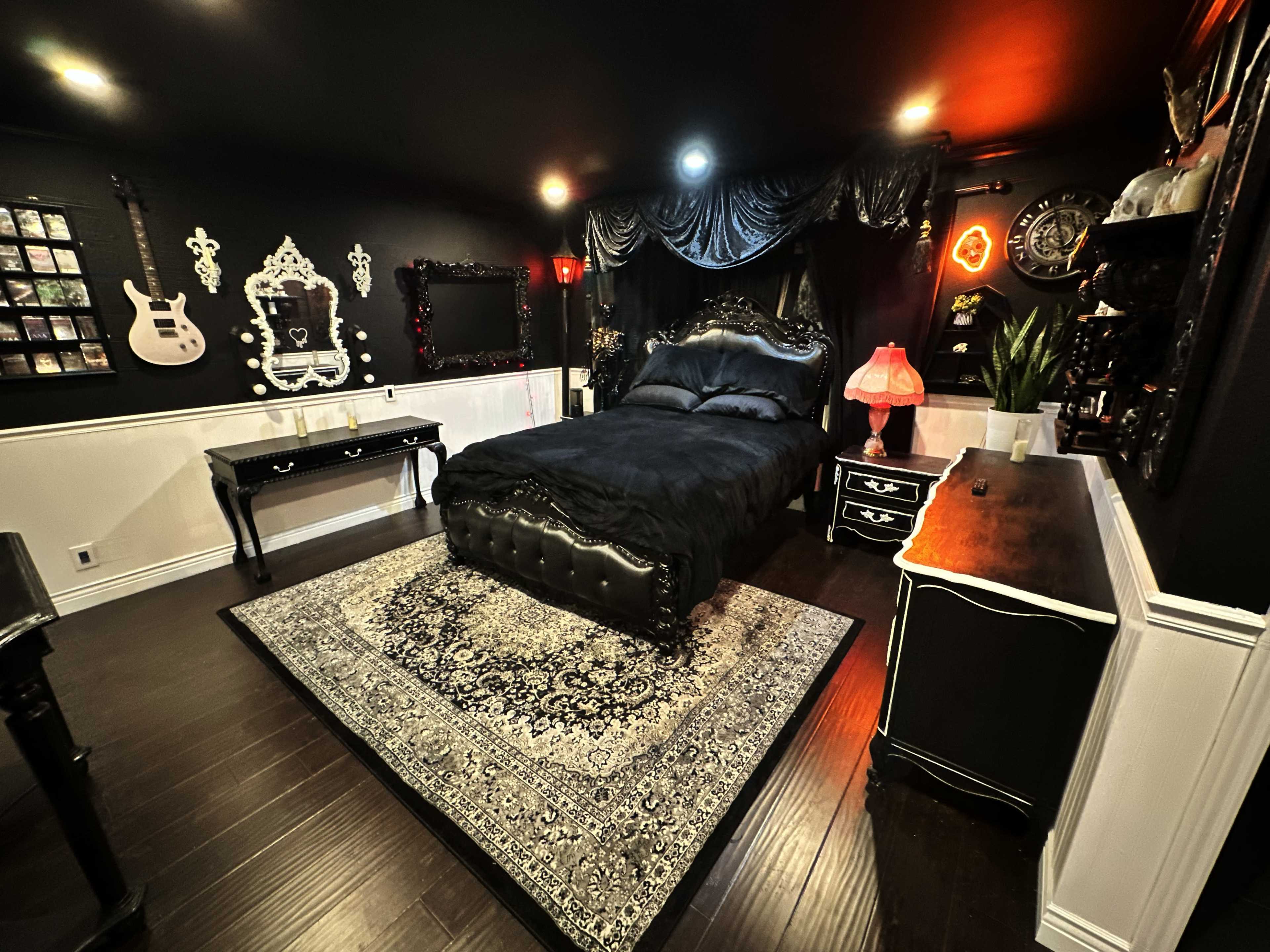 The image depicts a dark-themed bedroom featuring a black ornate bed with a decorative headboard, a patterned rug, and various furnishings including a dresser and side tables.