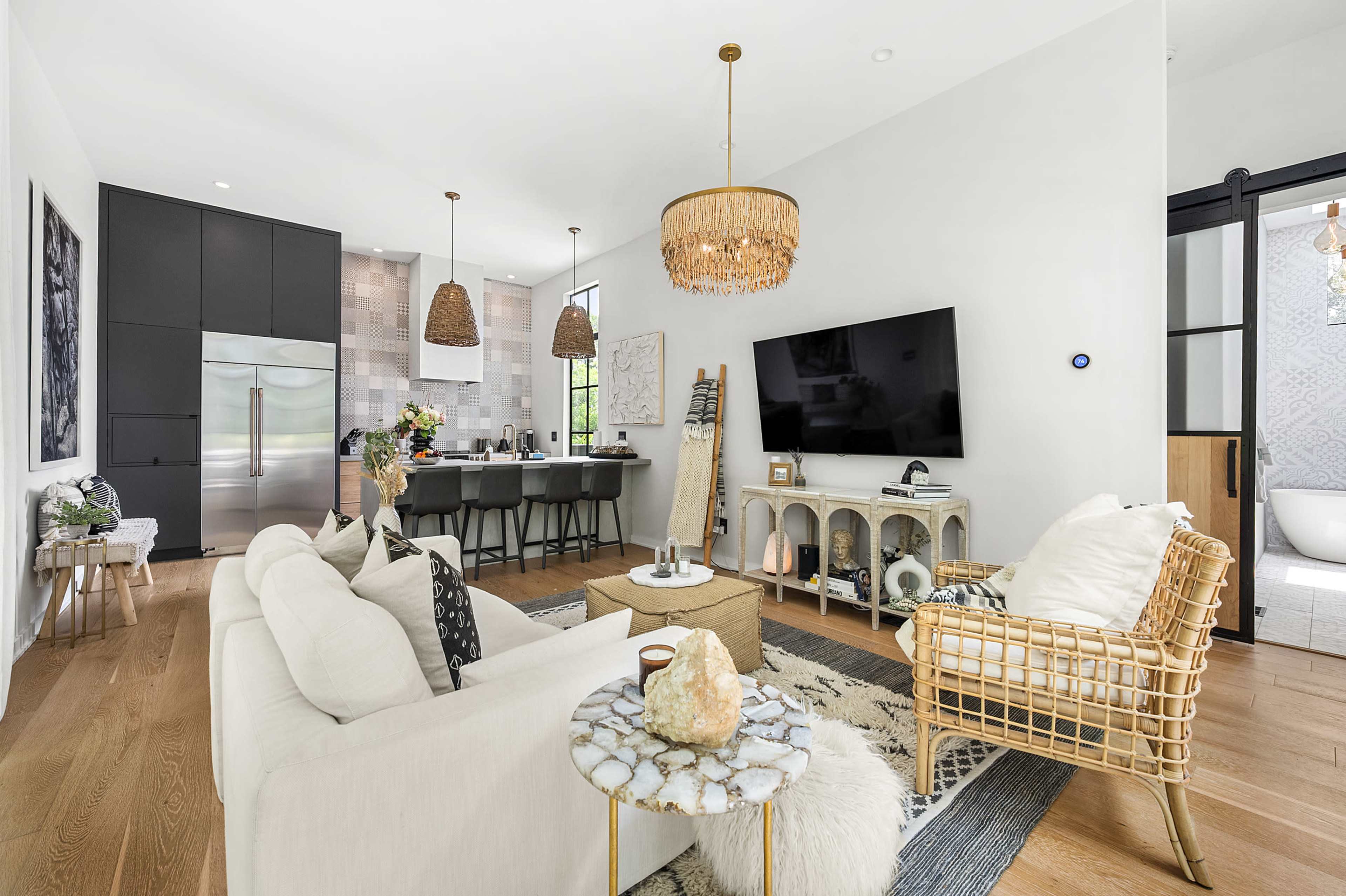 A modern living room features a beige sofa, a rattan chair, a large TV on the wall, and a kitchen area with bar stools in the background.