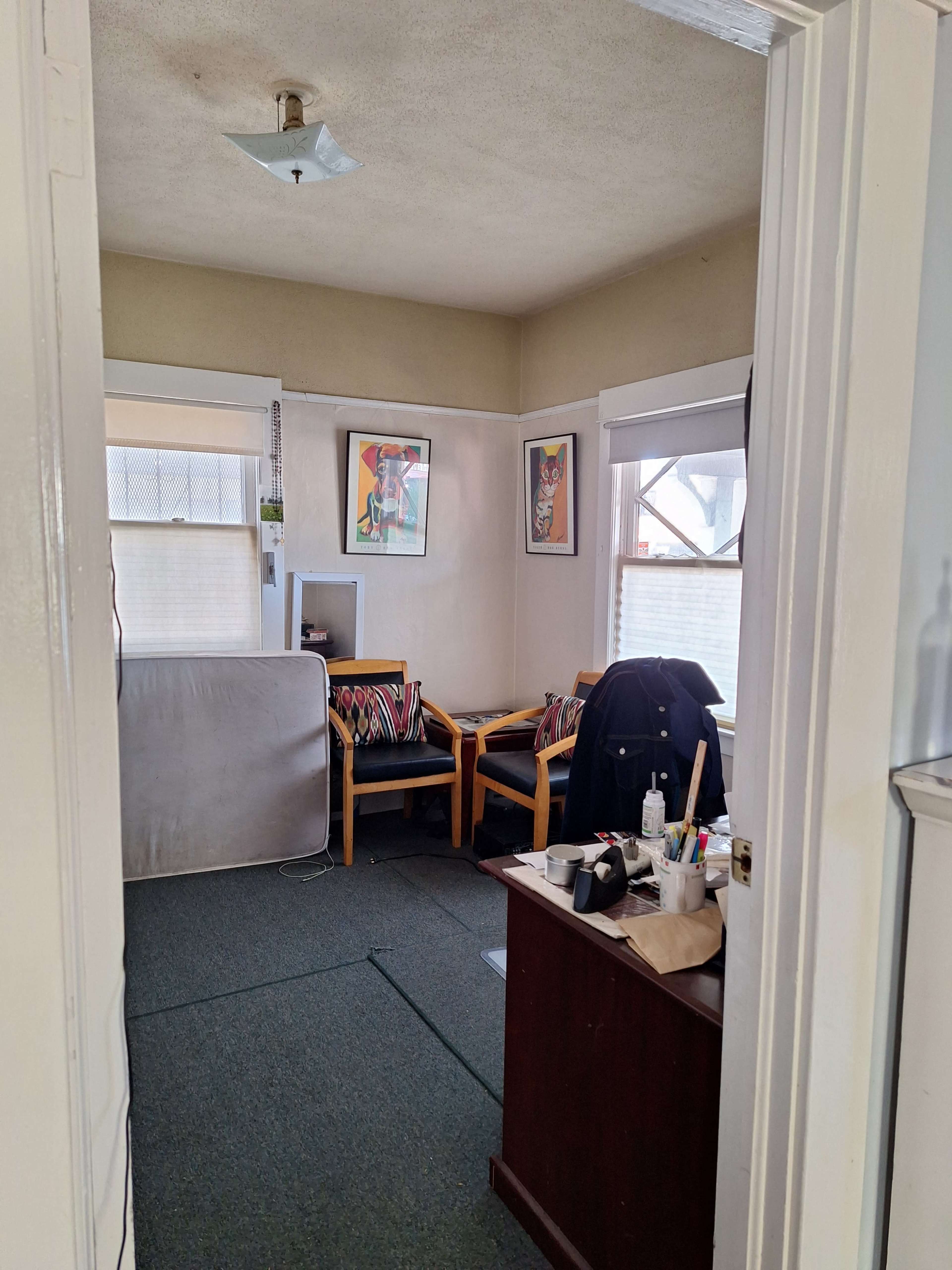 The image shows a small room with two wooden chairs, a folding mattress leaning against the wall, and a desk with various items on it.