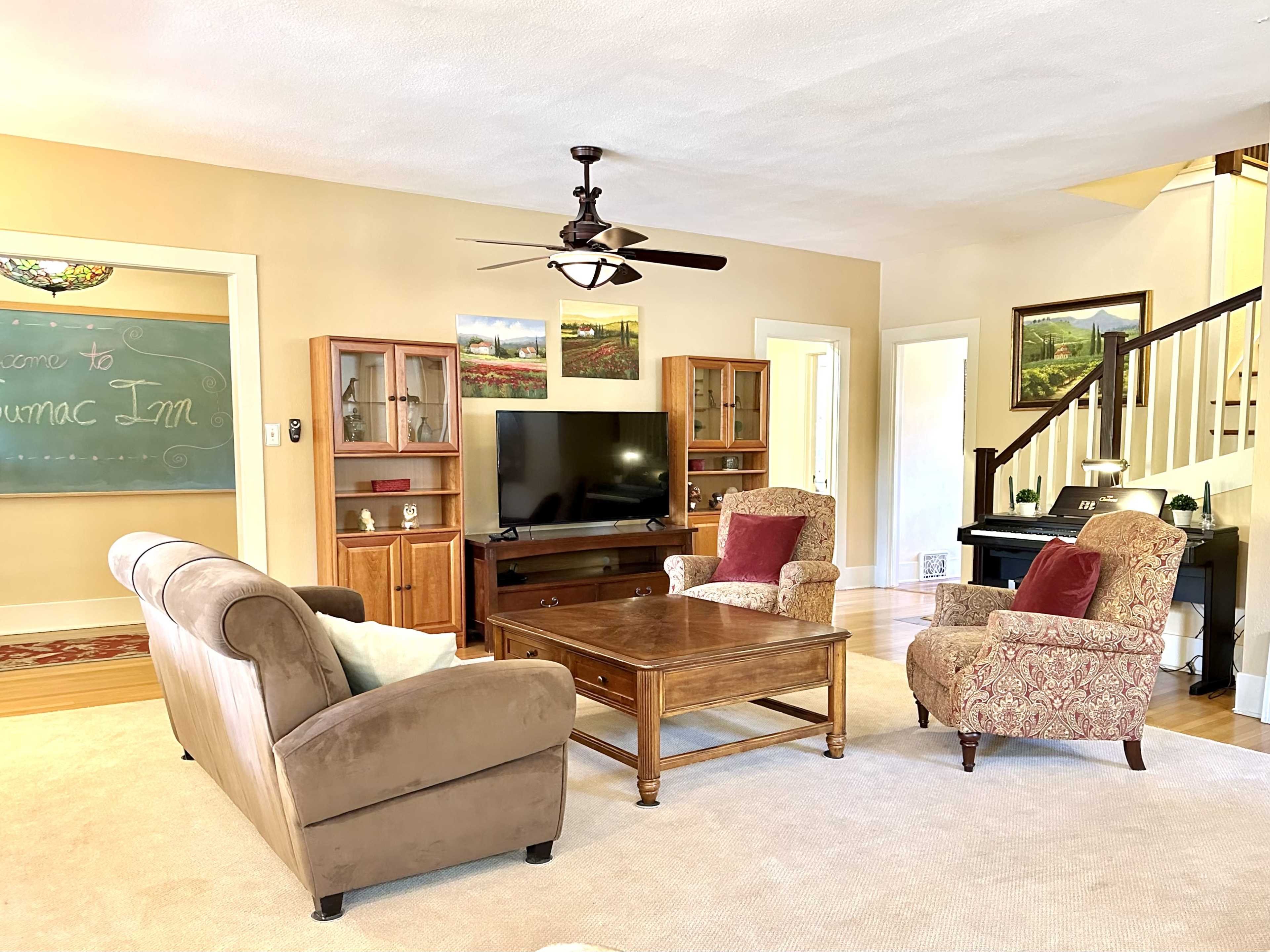 The image shows a cozy living room with a brown sofa, two upholstered chairs, a wooden coffee table, a television, and a staircase in the background.