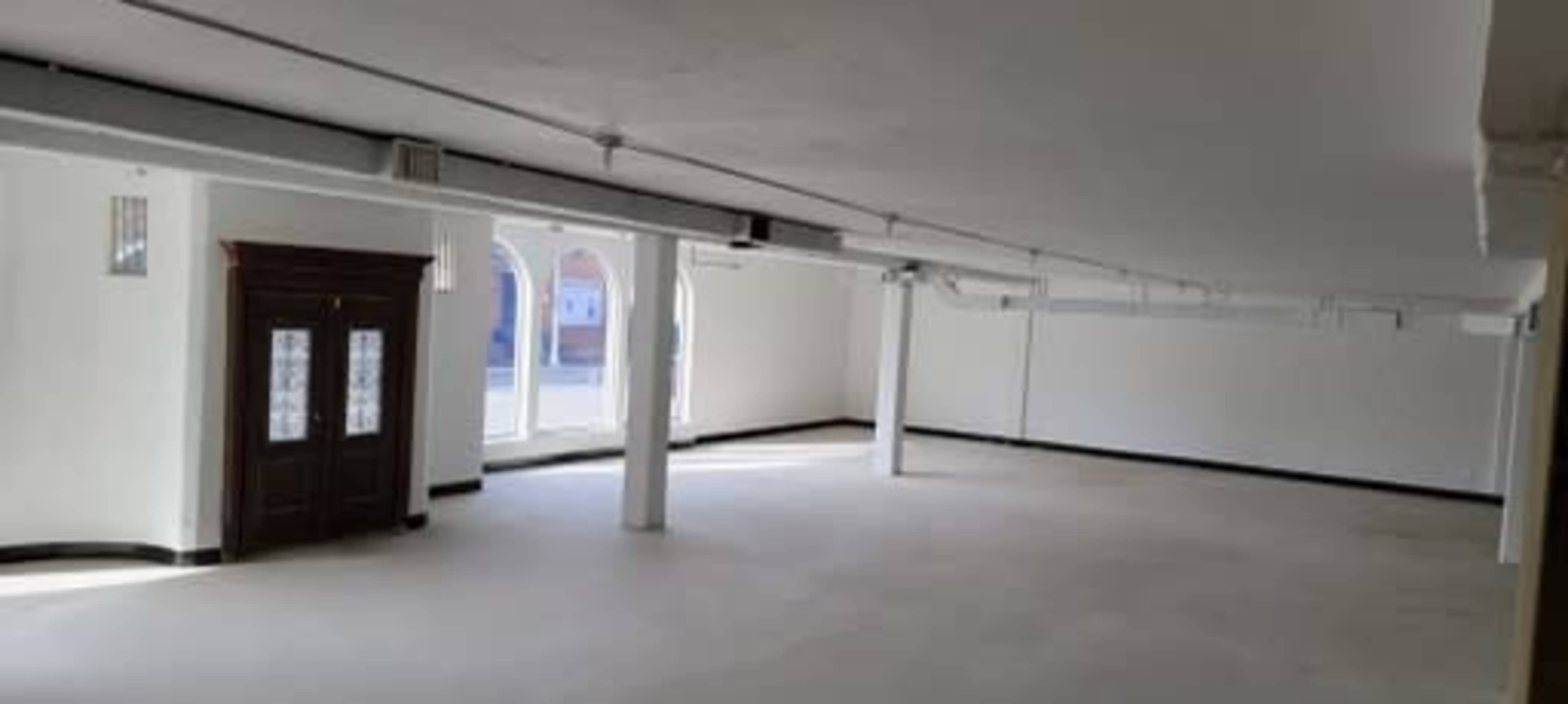 A large, empty commercial space with white walls, concrete flooring, and tall columns, featuring a wooden cabinet against one wall.