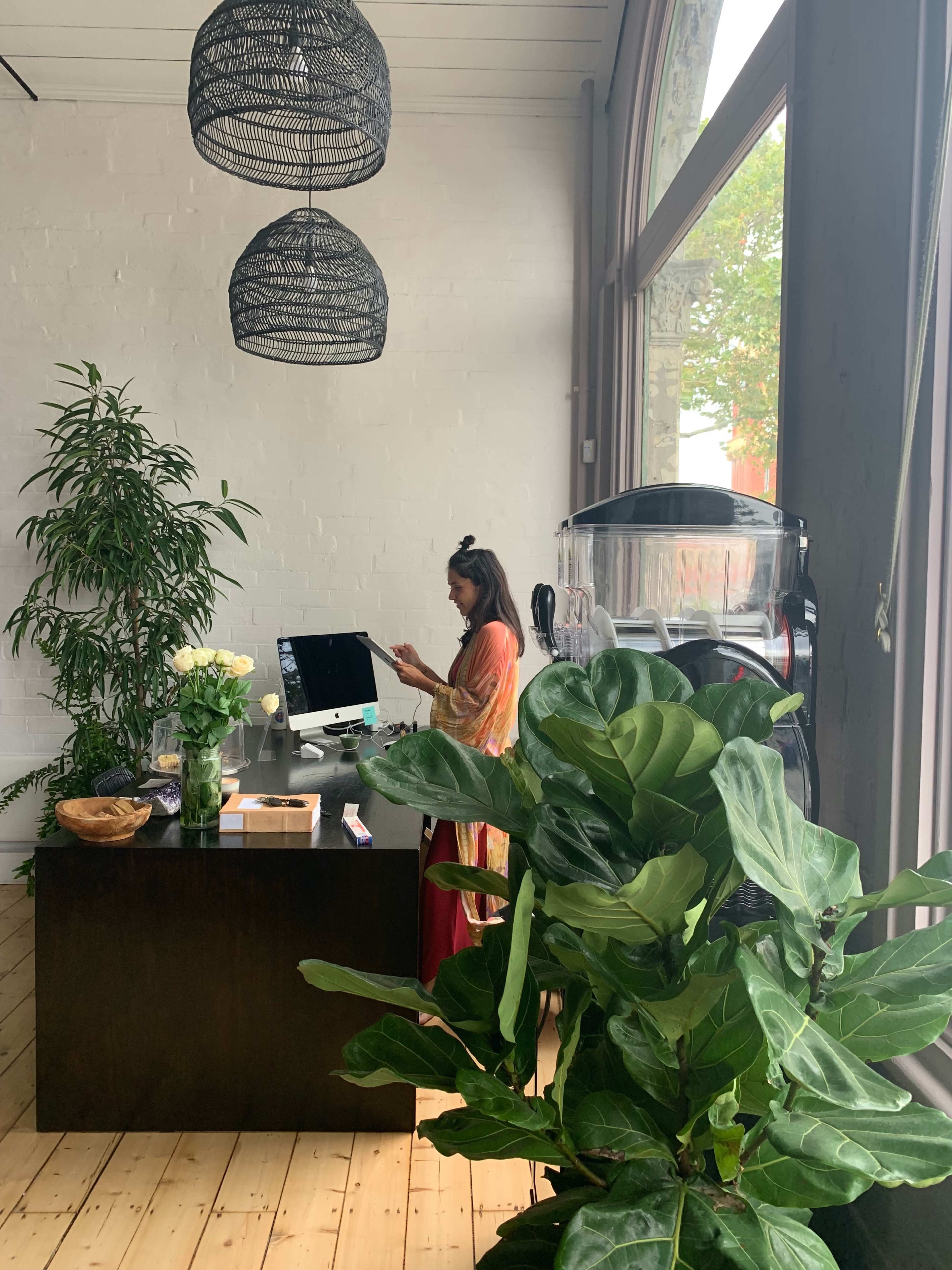 A woman stands at a desk with a computer in a bright, plant-filled room featuring large windows and decorative light fixtures.