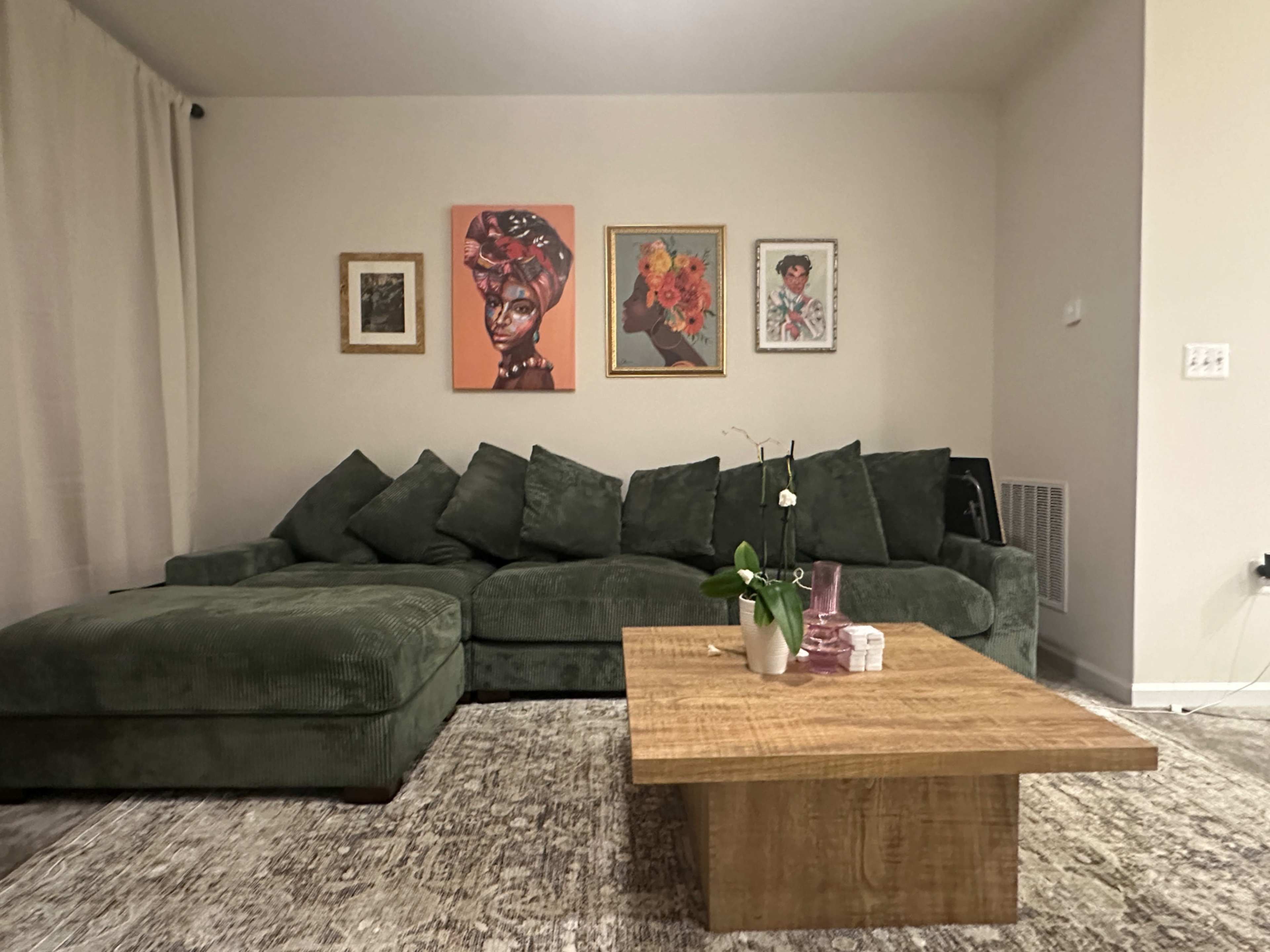 The image shows a living room with a green sectional sofa, a wood coffee table, and several framed artworks on the walls.