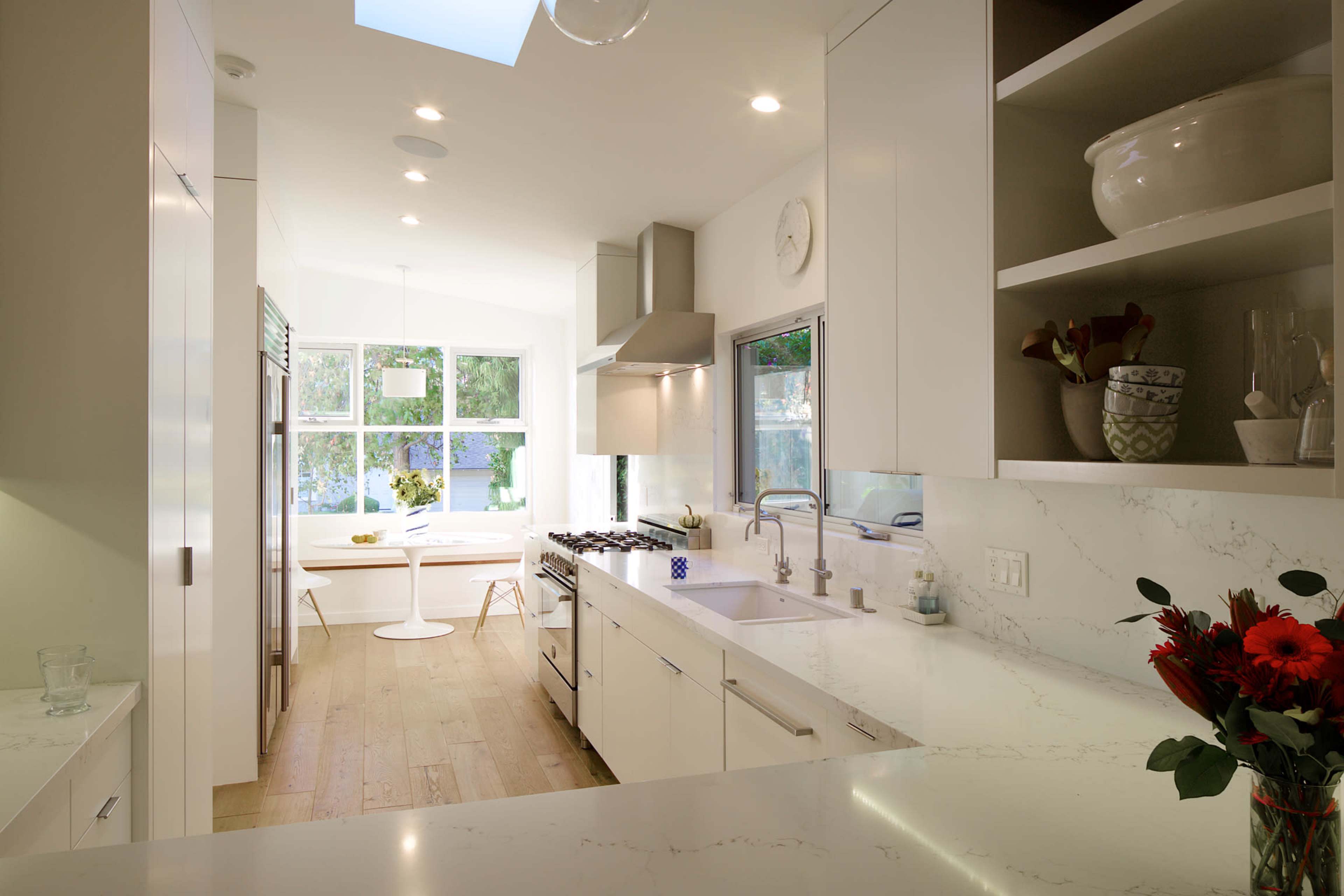 A sleek, modern kitchen features white cabinetry, a marble countertop, and a view of a sunlit dining area through large windows.