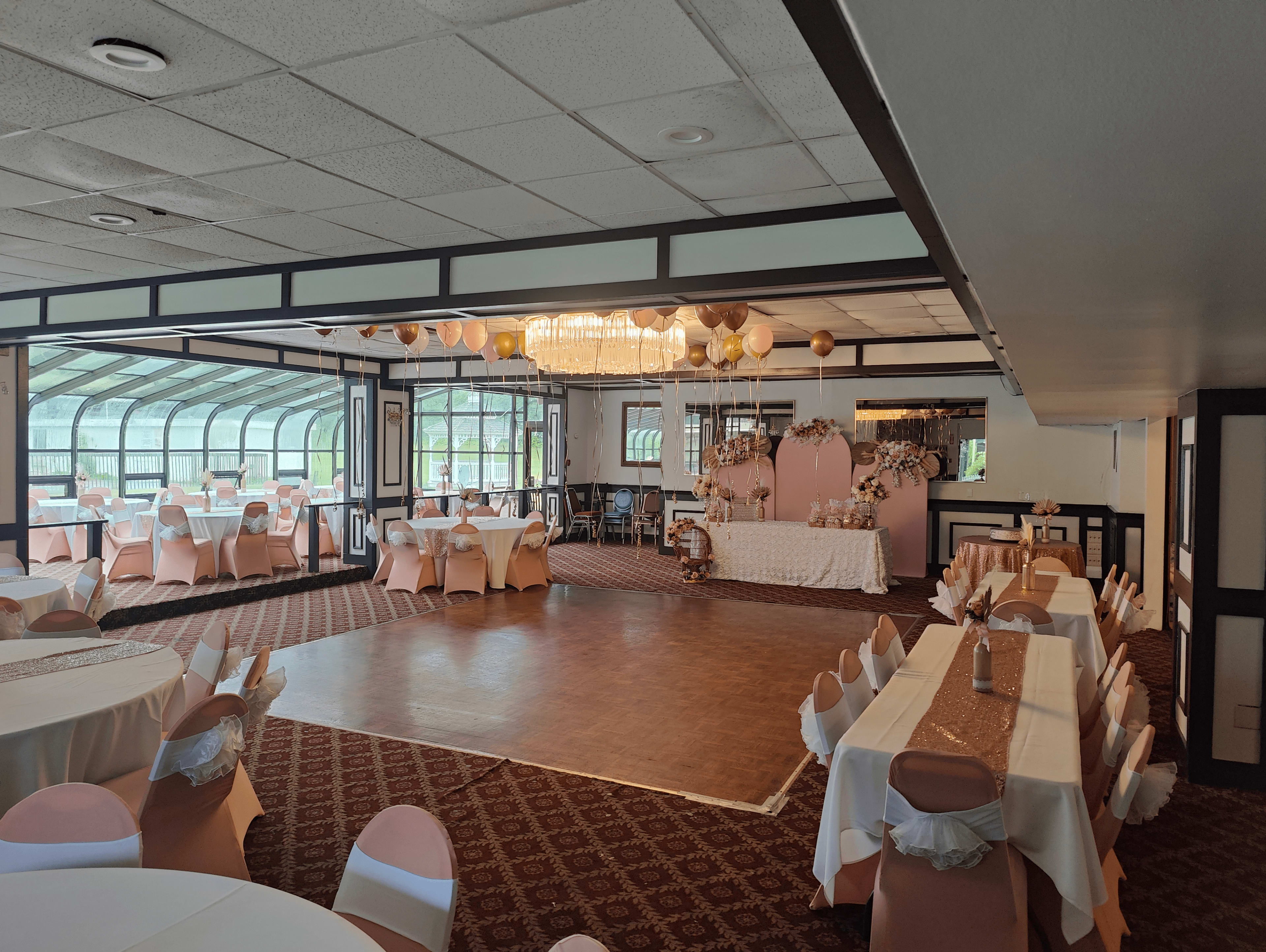 A banquet hall with round tables and chairs arranged around a central dance floor, featuring a decorated backdrop at one end.