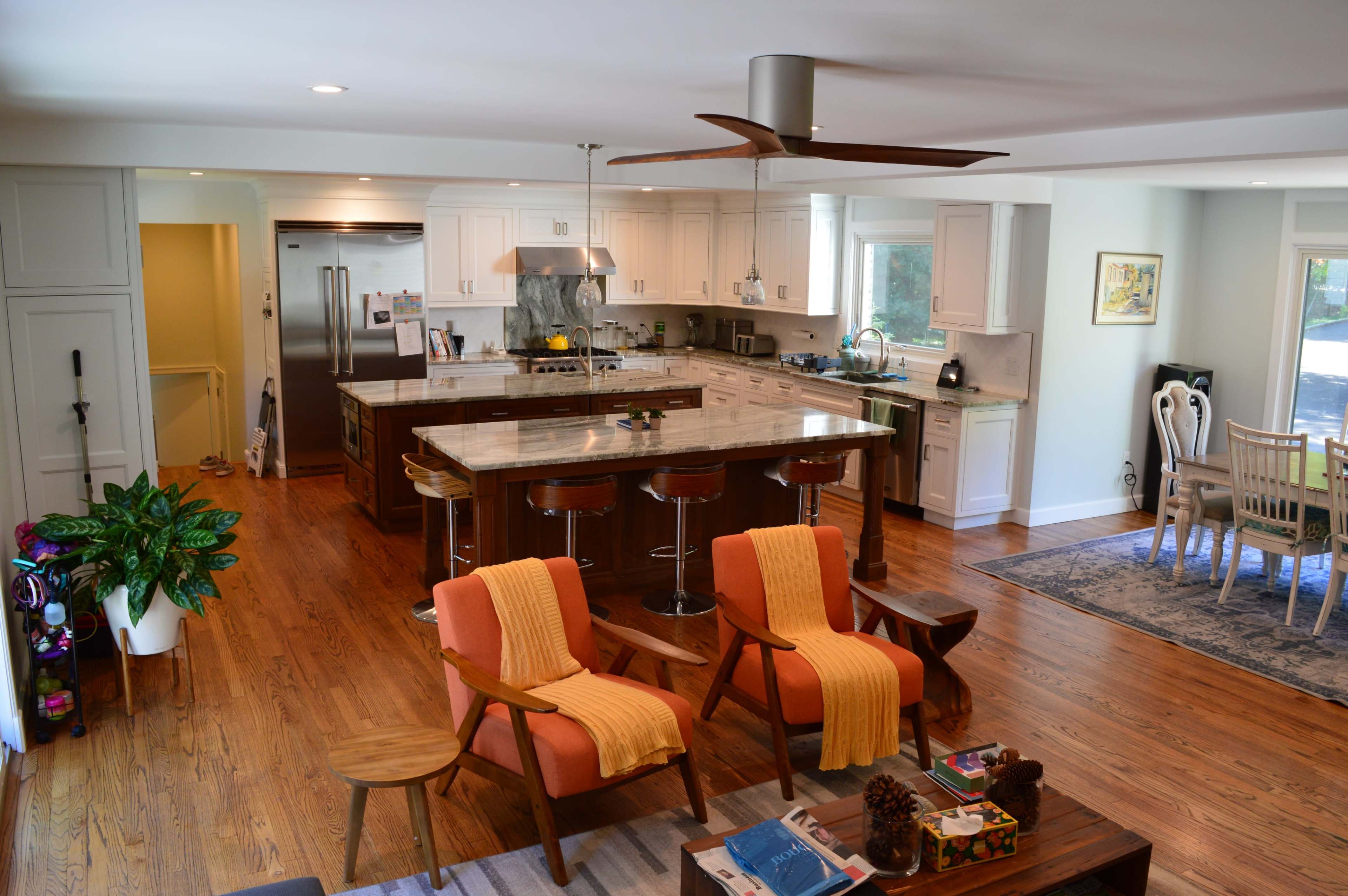 The image shows a bright and spacious kitchen and dining area with wooden floors, an island with bar stools, and comfortable seating with orange throws.