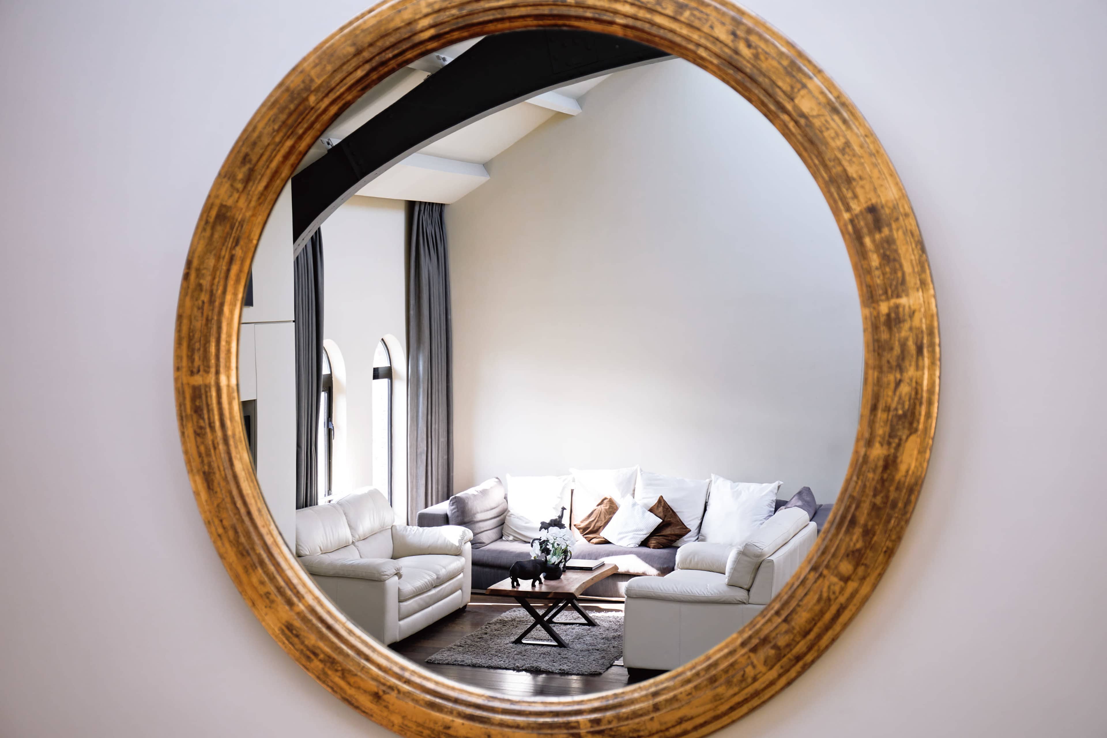 A round gold-framed mirror reflects a cozy living room with white sofas and a coffee table.