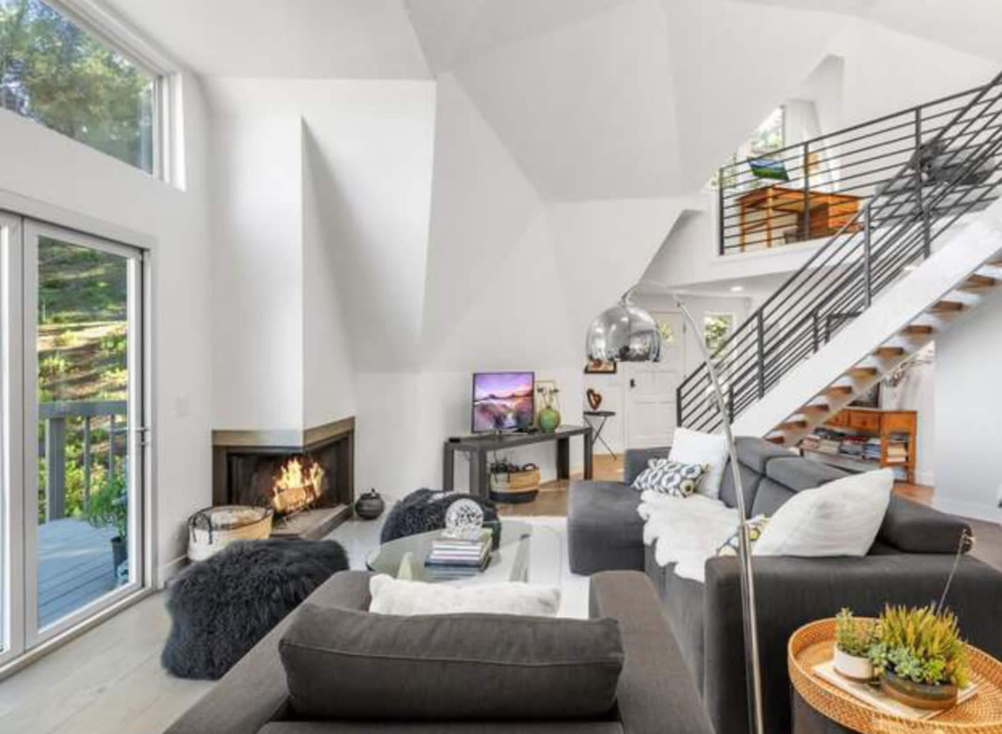 The image shows a modern living room with a high ceiling, featuring a fireplace, a staircase, and large windows allowing natural light to enter.