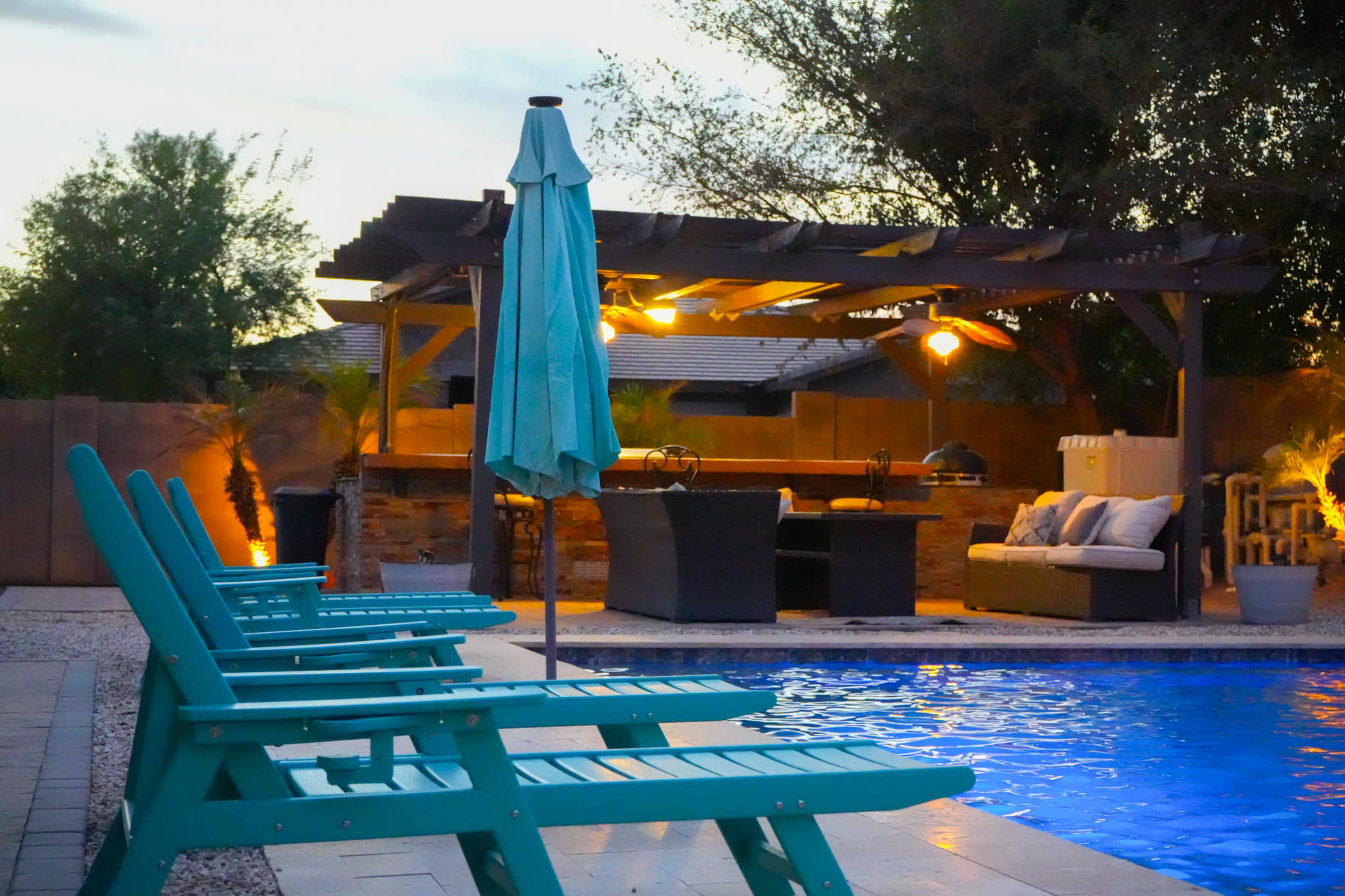 The image shows a backyard pool area with turquoise lounge chairs, a shaded seating area, and outdoor lighting.