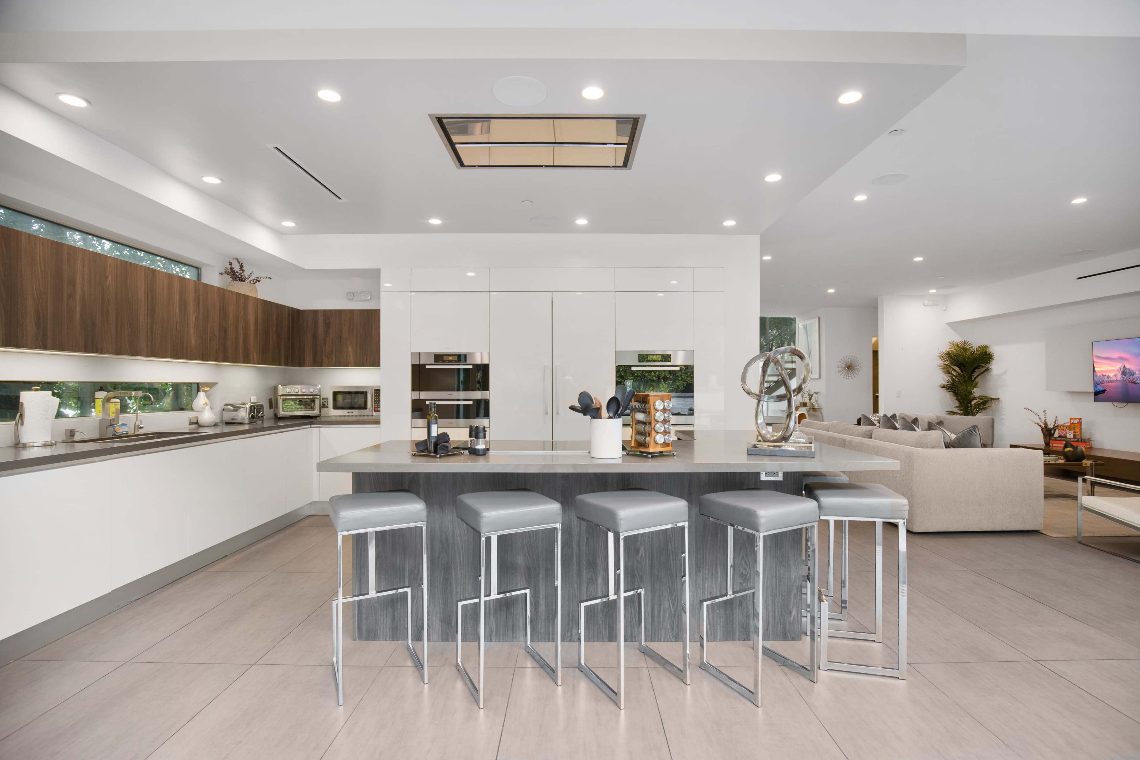 The image shows a modern kitchen with a large island, featuring stainless steel bar stools, sleek cabinetry, and integrated appliances.