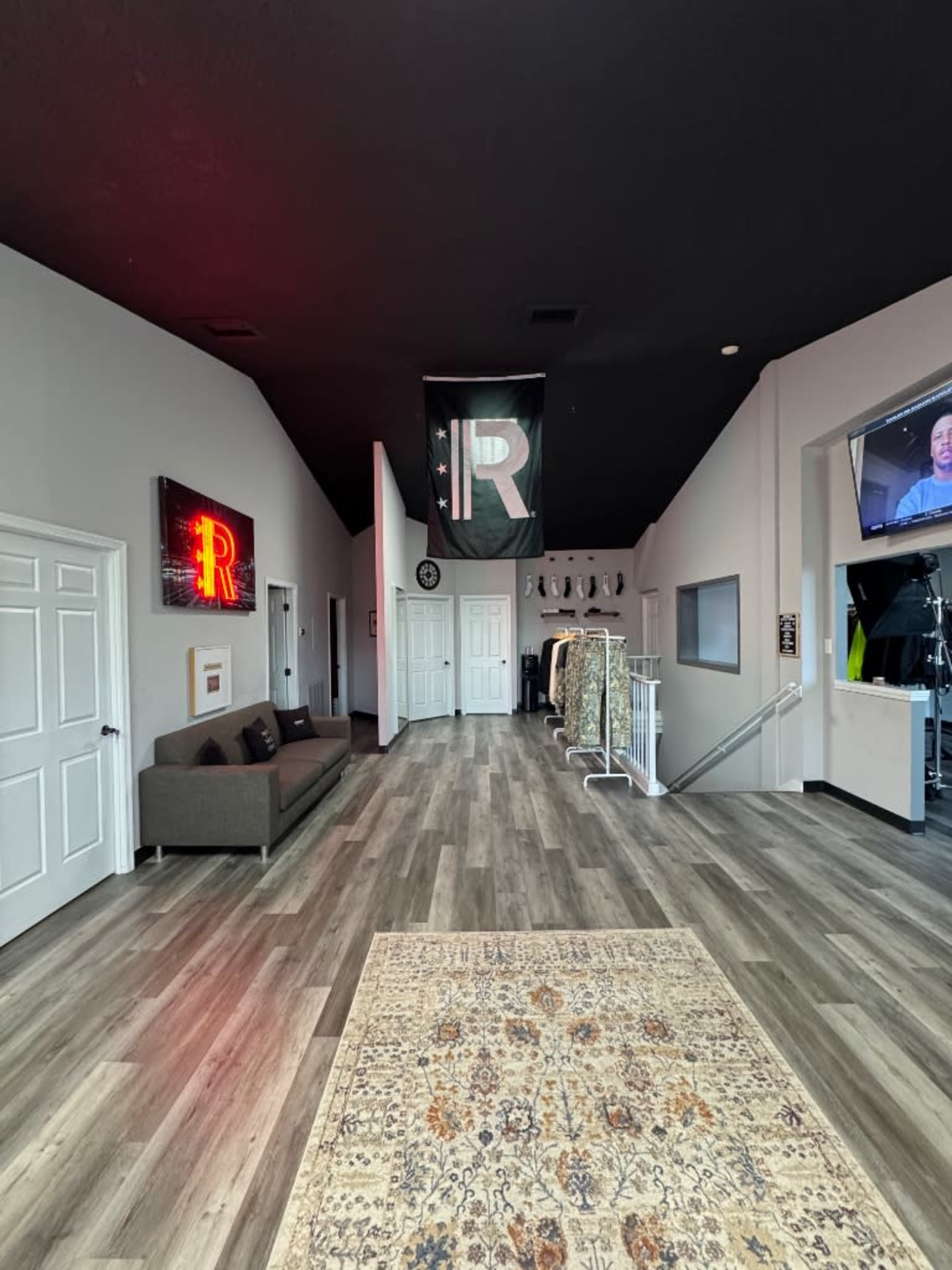 The image shows a modern interior hallway with a decorative rug, a couch, and a large flag with the letter "R" hanging on the wall, alongside various clothing items displayed.