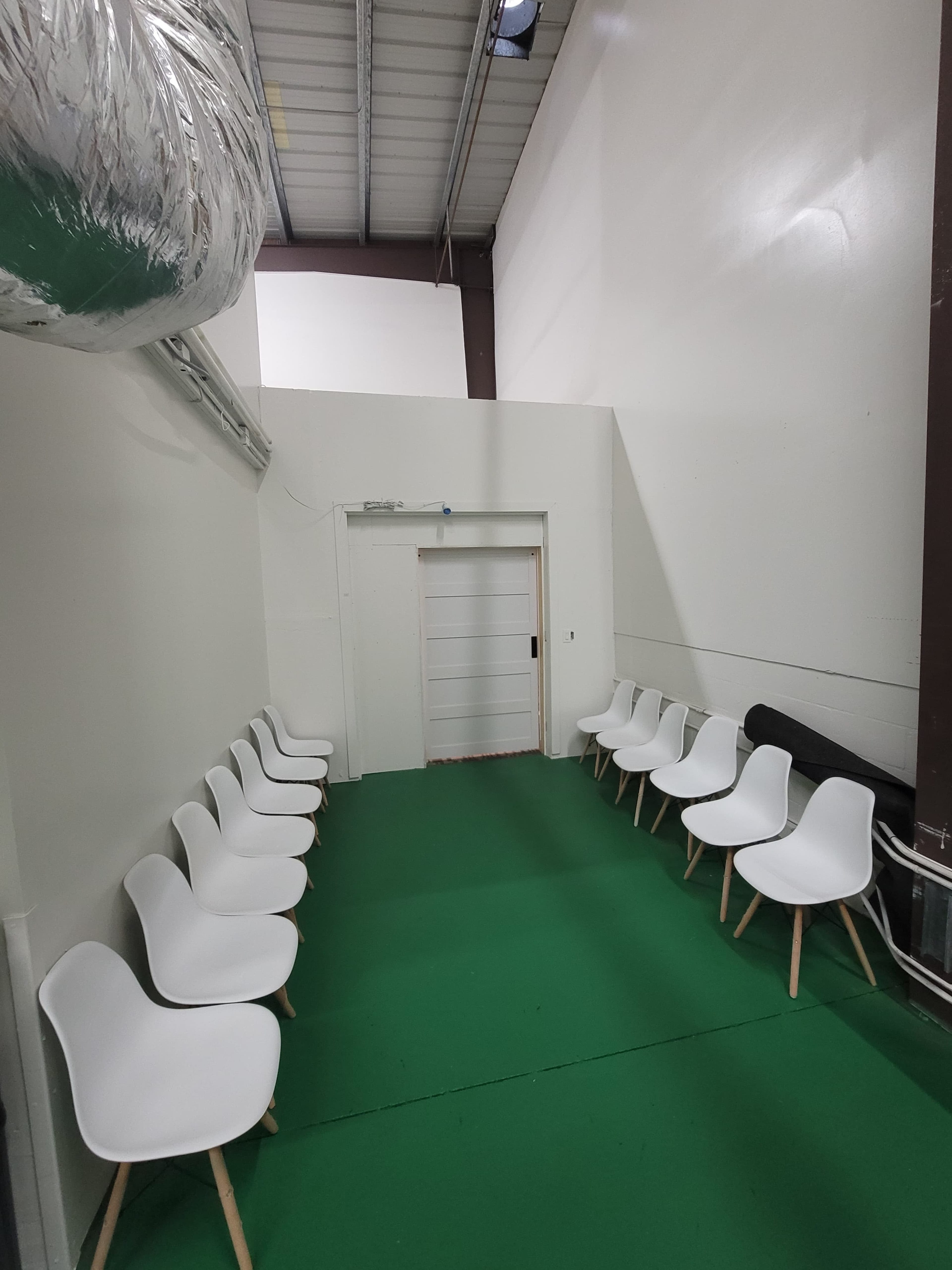 The image shows a sparsely furnished waiting area with white chairs arranged neatly along the walls and a green floor.