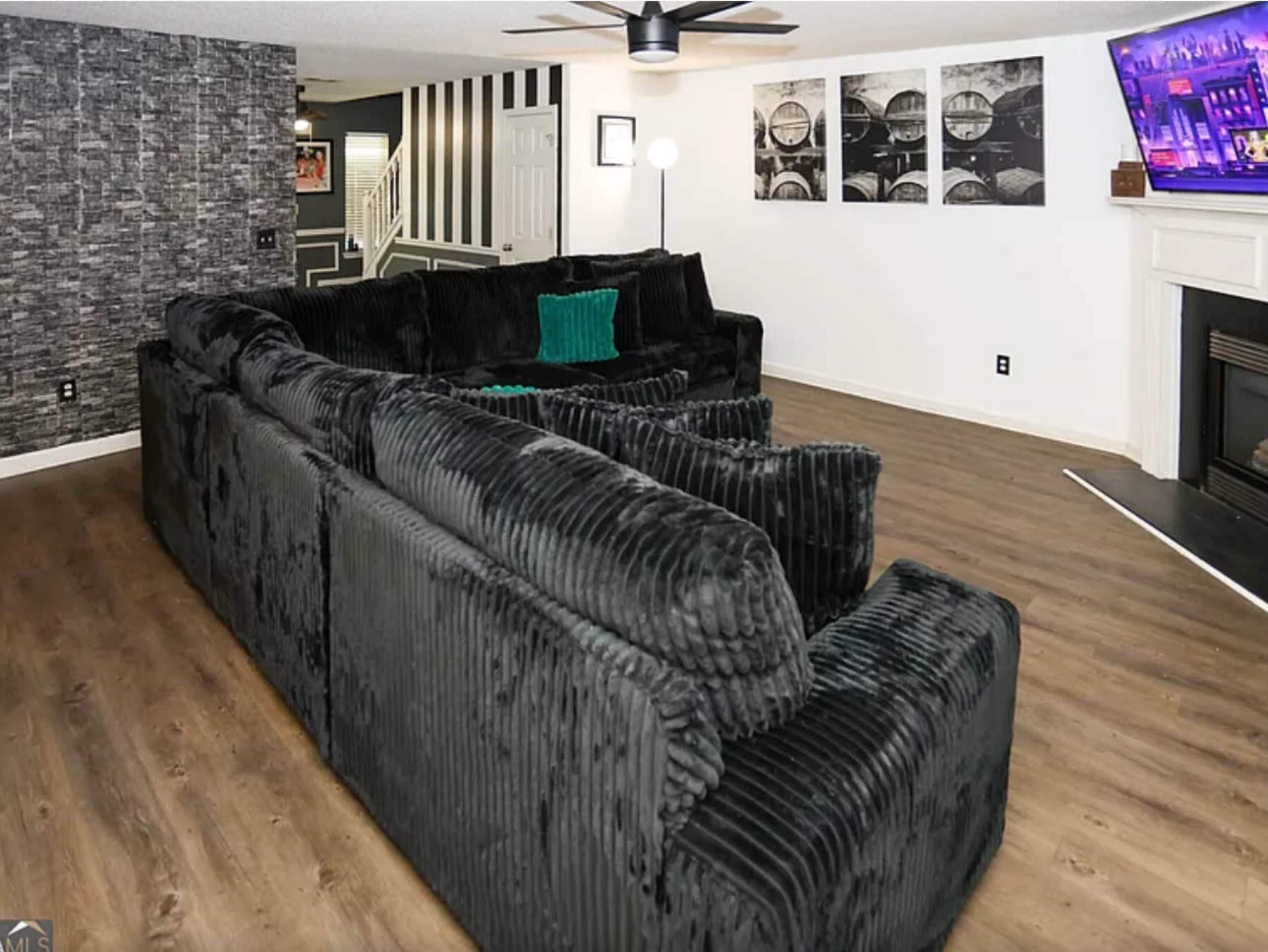 The image shows a spacious living area featuring a large, dark-colored sectional sofa, a flat-screen TV mounted on the wall, and a fireplace in the corner.