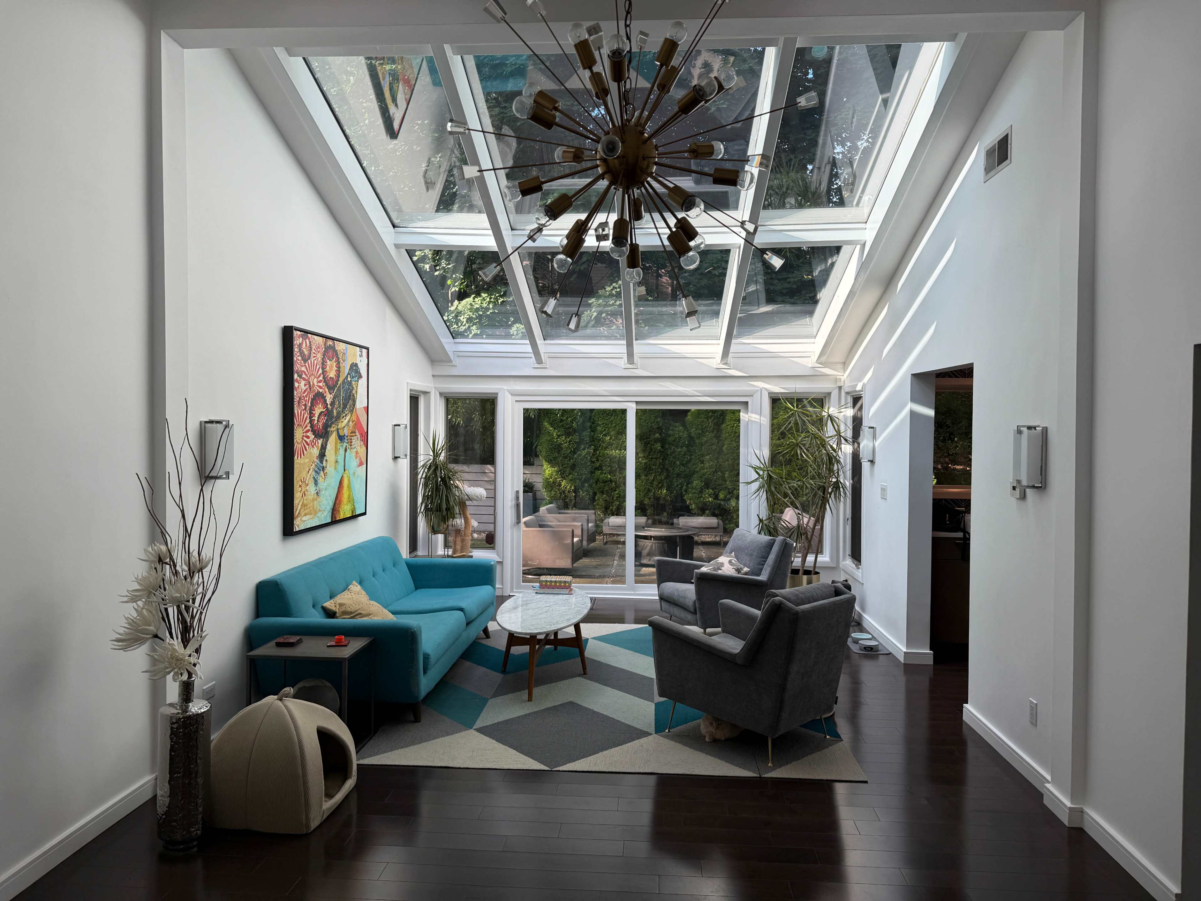 The image shows a modern living room featuring a turquoise sofa, a gray armchair, a geometric-patterned rug, and large windows overhead that allow natural light to flood the space.