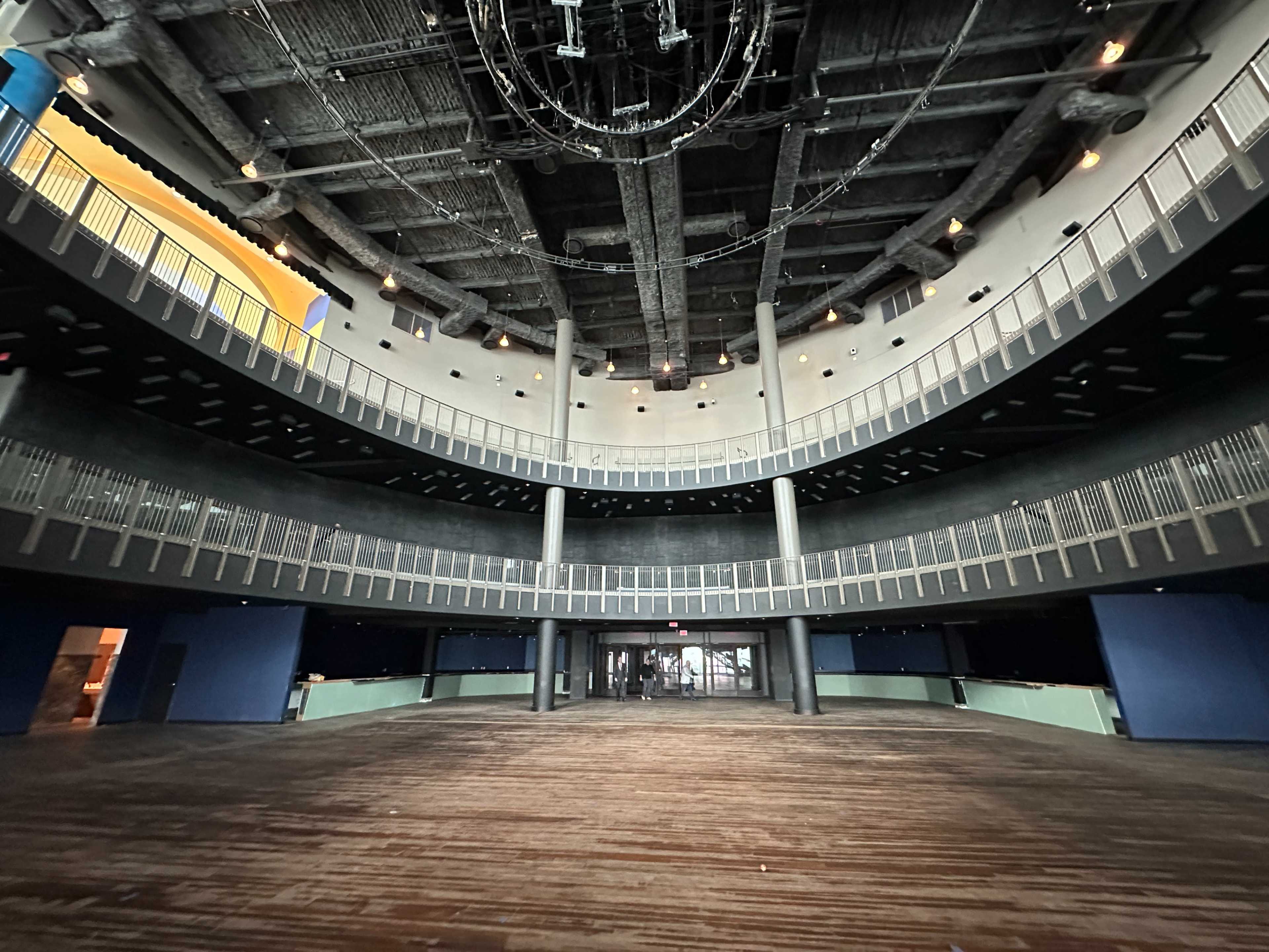 A spacious indoor auditorium with multi-level seating, a wooden floor, and large windows at the entrance.