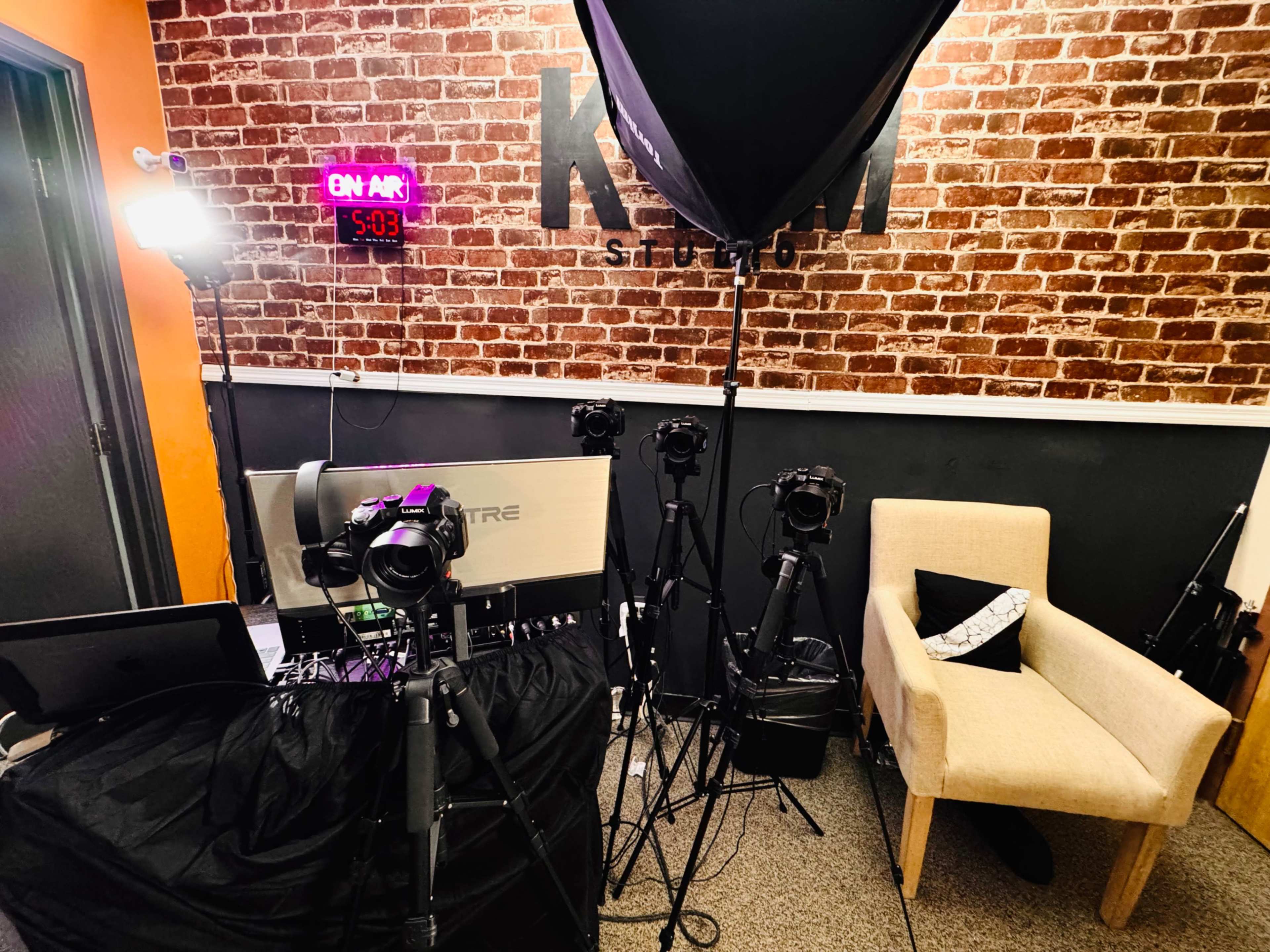 The image shows a photography studio with multiple cameras on tripods, a large softbox light, and a comfy chair against a brick wall.
