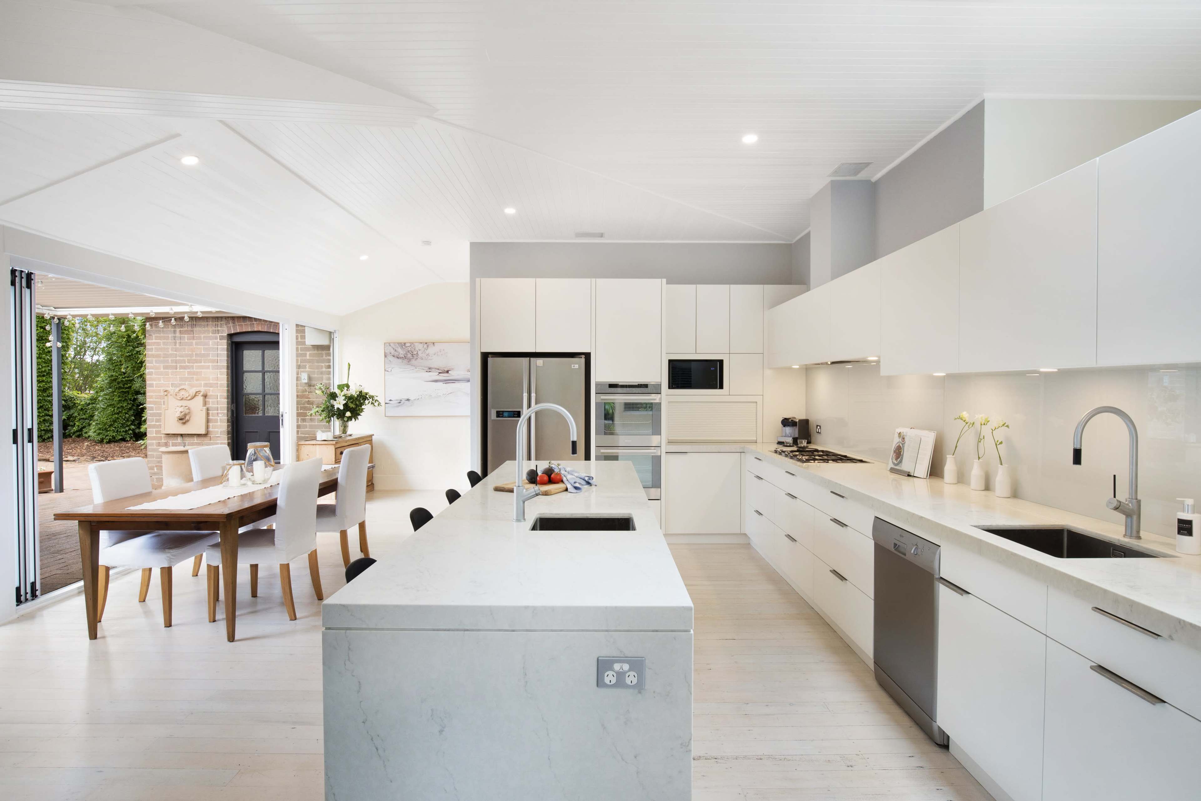 A modern kitchen featuring white cabinetry, stainless steel appliances, and an island with a double sink, adjacent to a dining area with a wooden table.