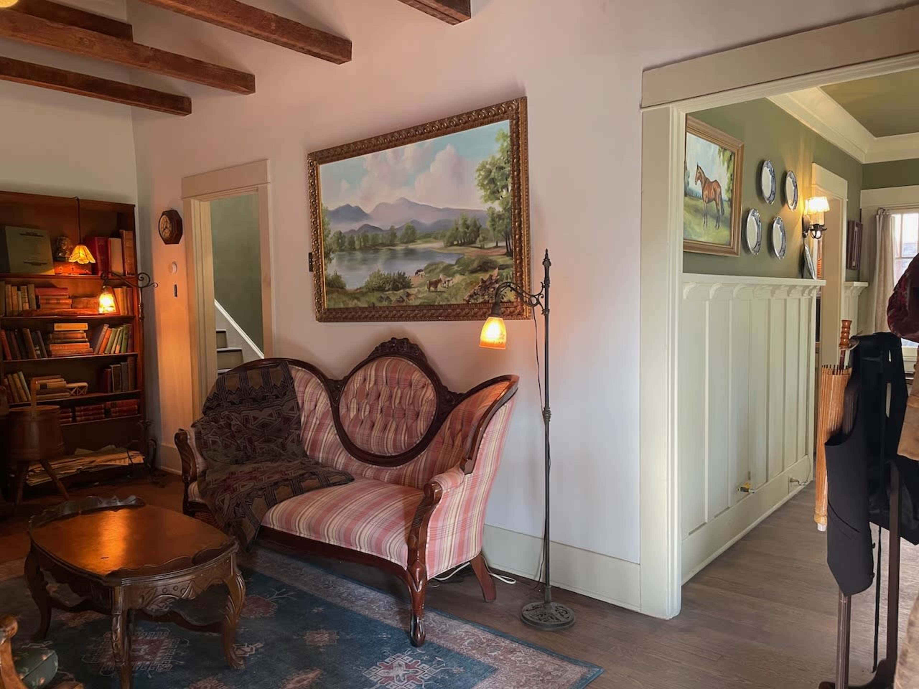 The scene features a traditional living room with a vintage pink-striped sofa, a decorative painting of a landscape, and a lamp beside the sofa, all set against a backdrop of wooden beams and light-colored walls.