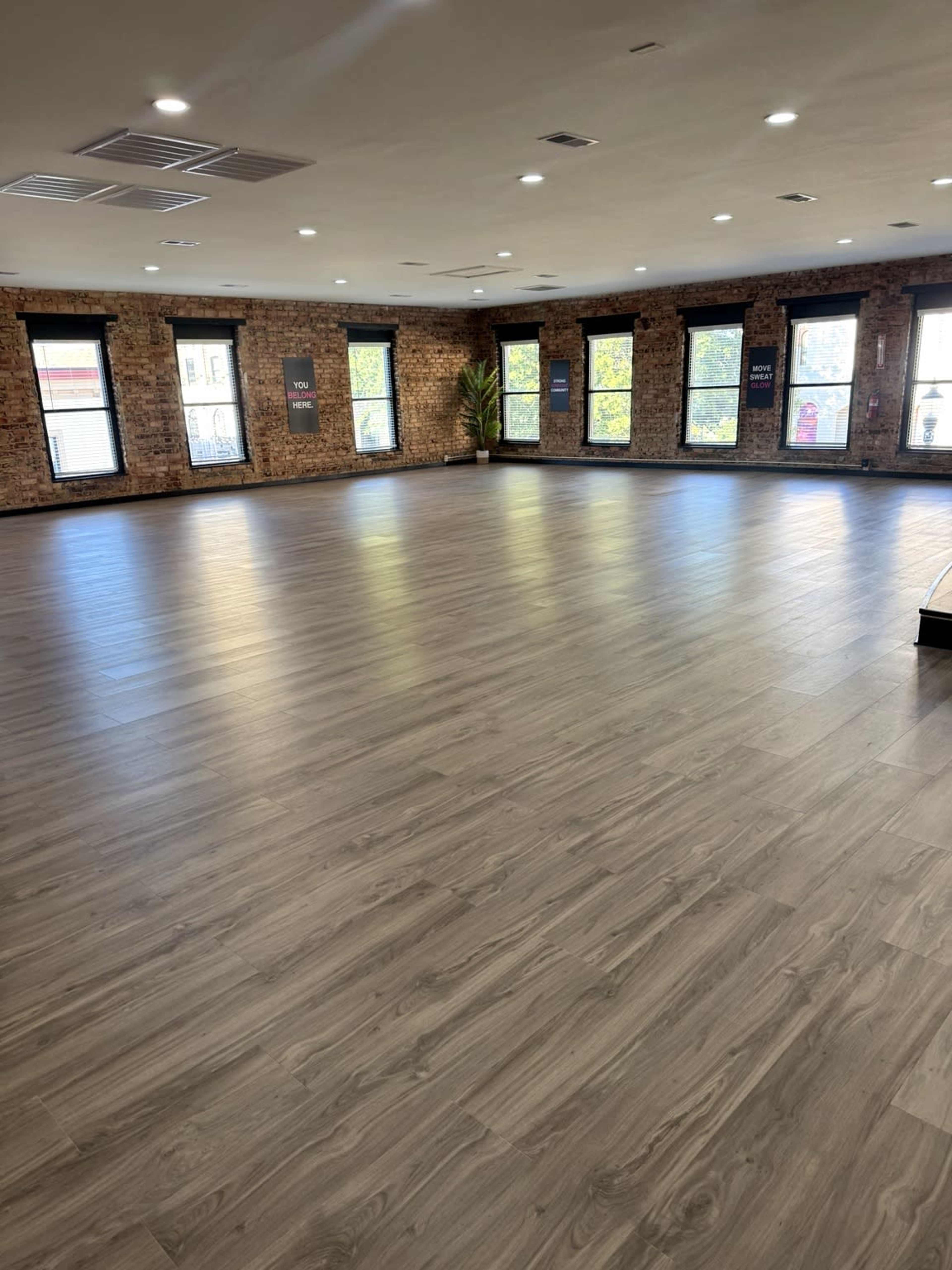 The image shows a spacious room with hardwood floors and large windows allowing natural light to enter.