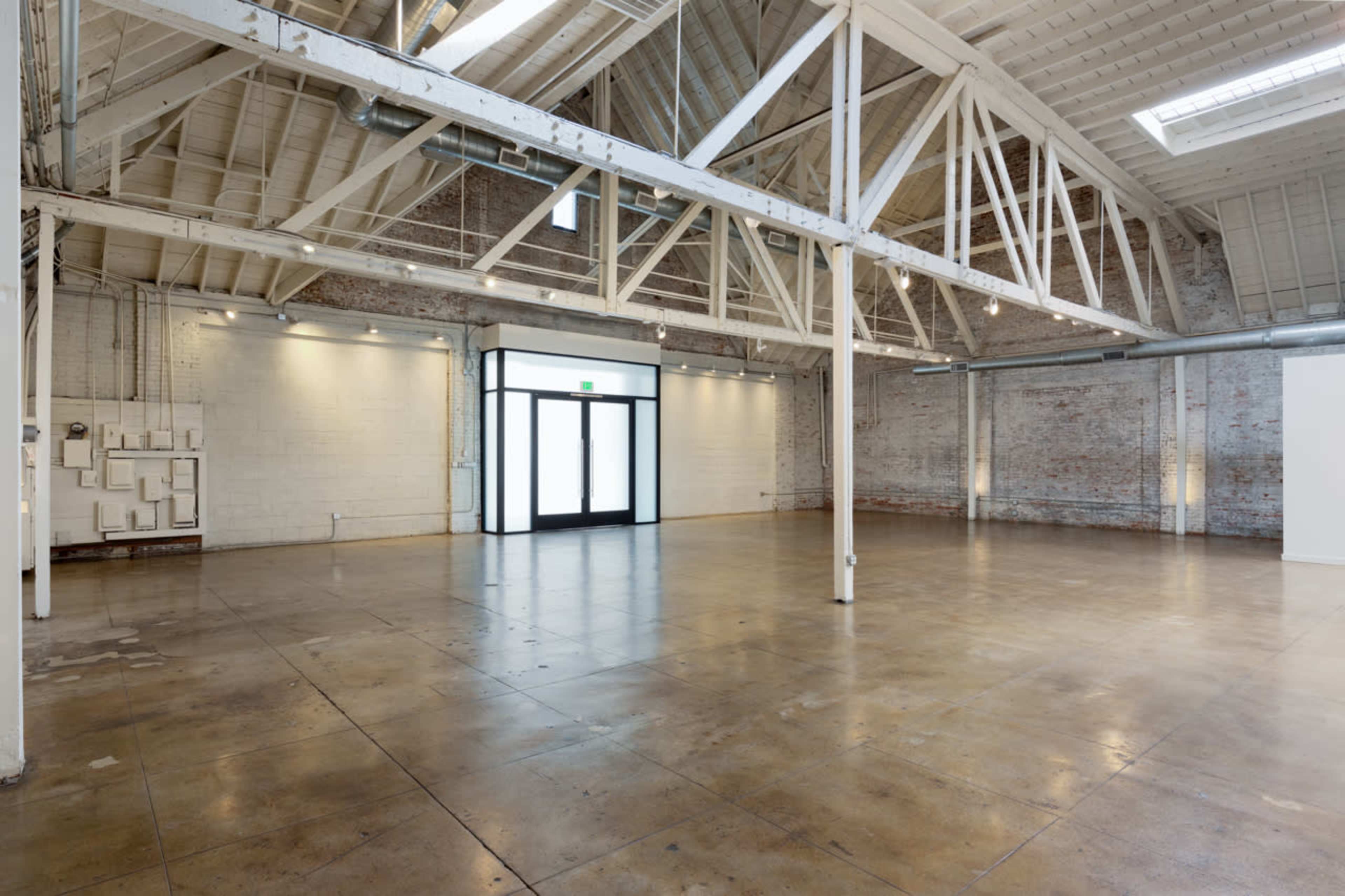 The image shows a spacious, empty industrial-style interior with high ceilings and exposed beams, featuring polished concrete floors and large windows.