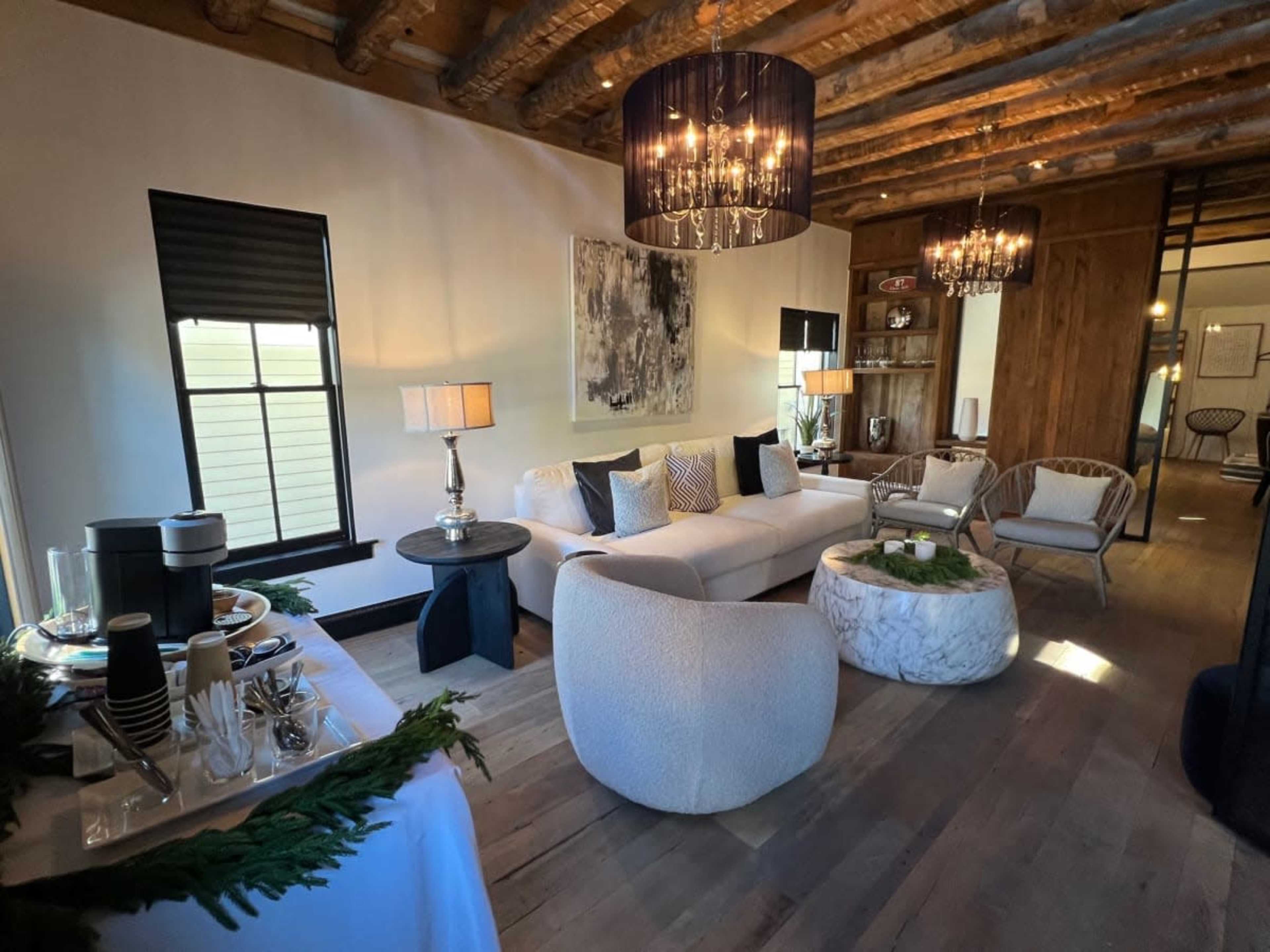 The image shows a modern living room with a white sectional sofa, a round marble coffee table, and several decorative lamps, all set against a backdrop of exposed wooden beams.