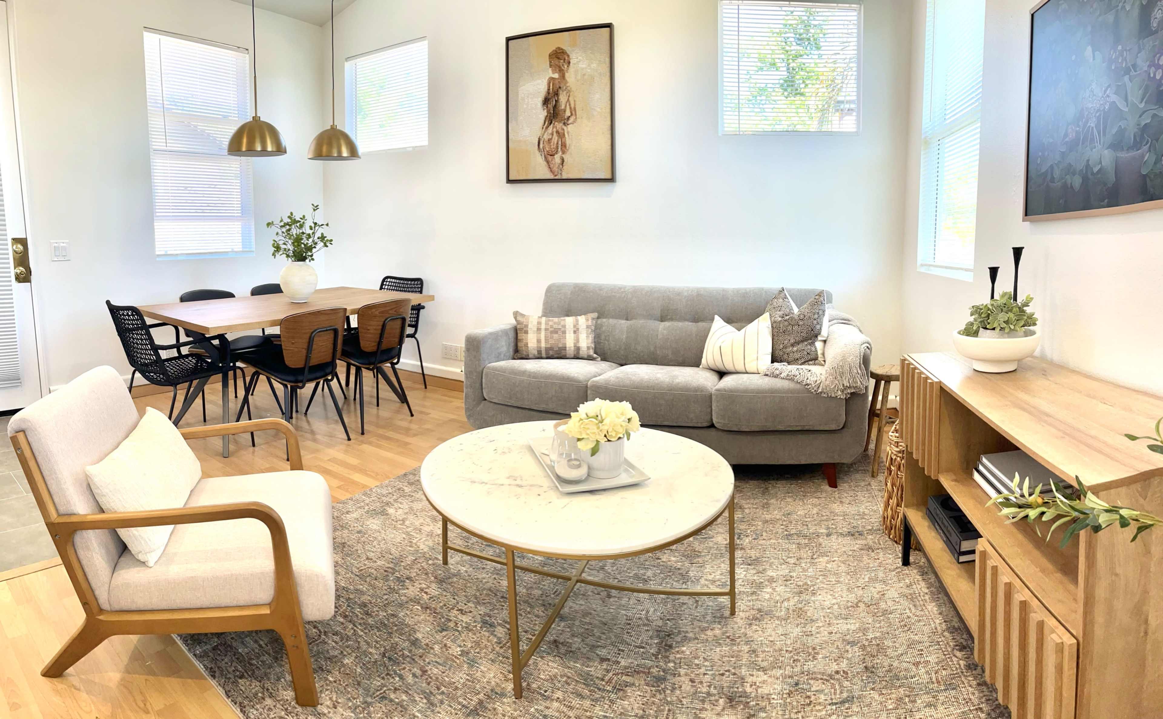 A modern living room with a gray sofa, a round marble coffee table, a wooden side table, and a dining area featuring a table and black chairs in a well-lit space.