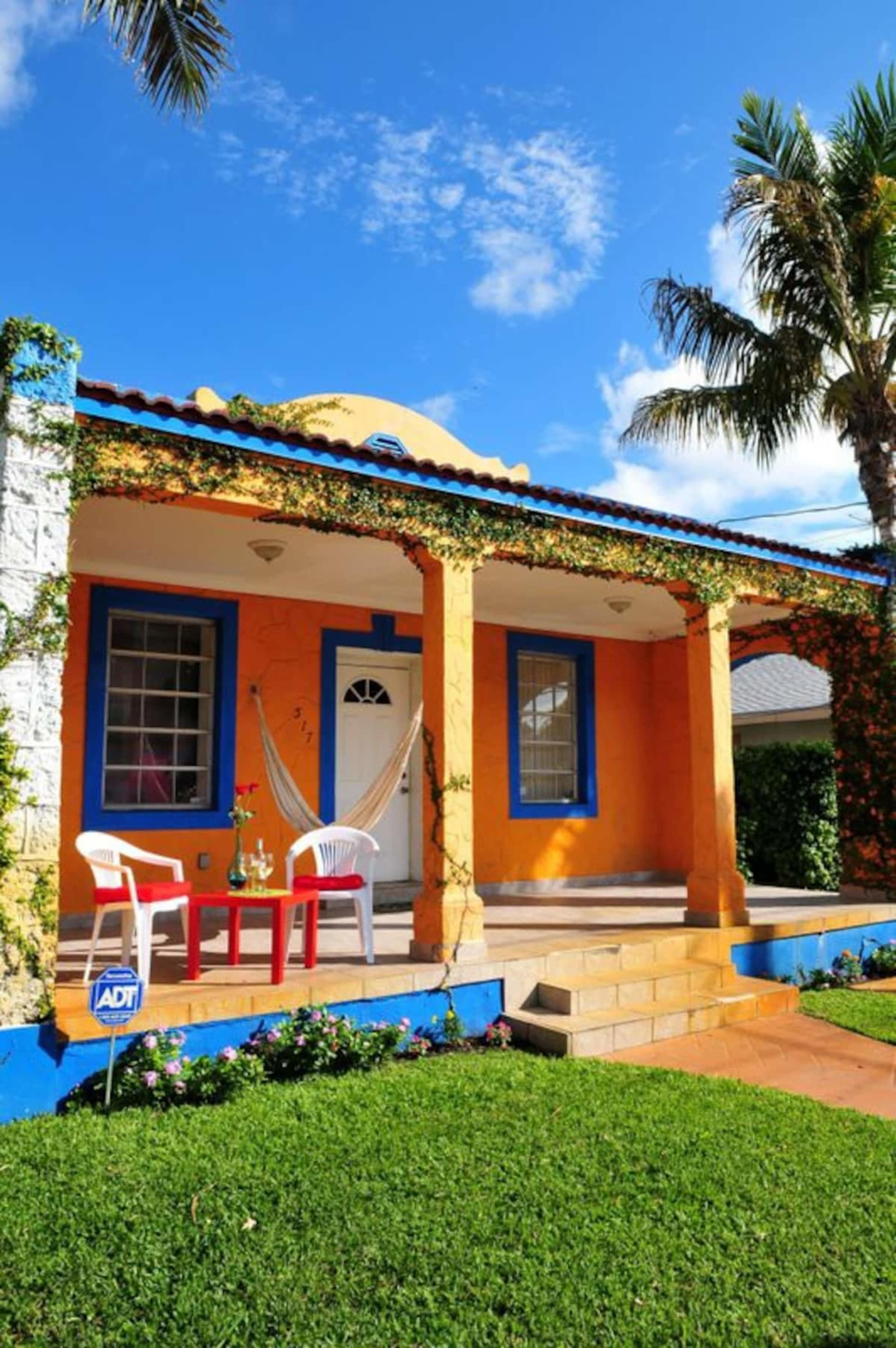 A colorful single-story house with a porch, surrounded by greenery and flowers, features two chairs and a table on the front step.