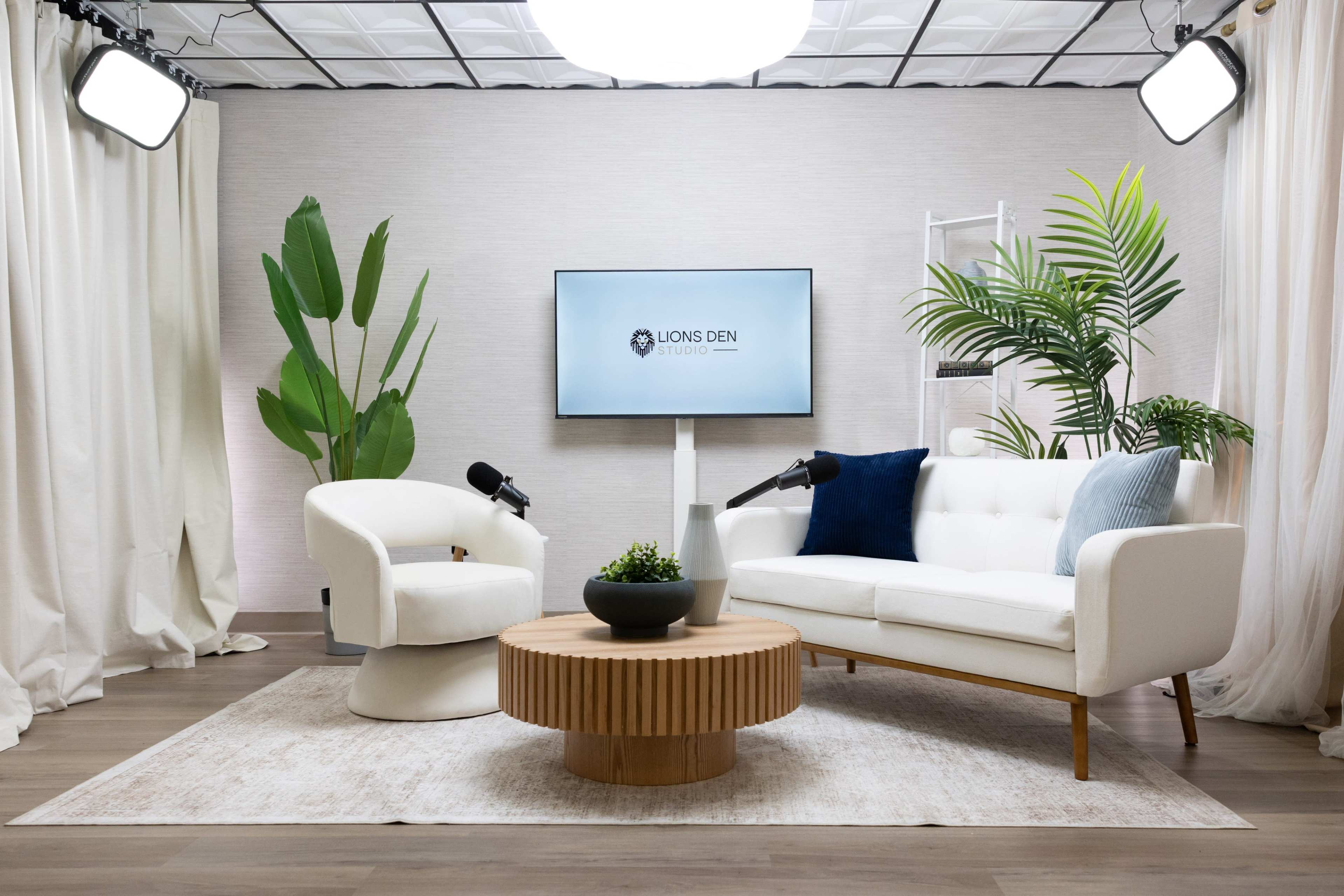 The image shows a modern studio setup with a white sofa, a round wooden coffee table, a large screen displaying "LIONS DEN," and decorative plants in a bright, well-lit space.