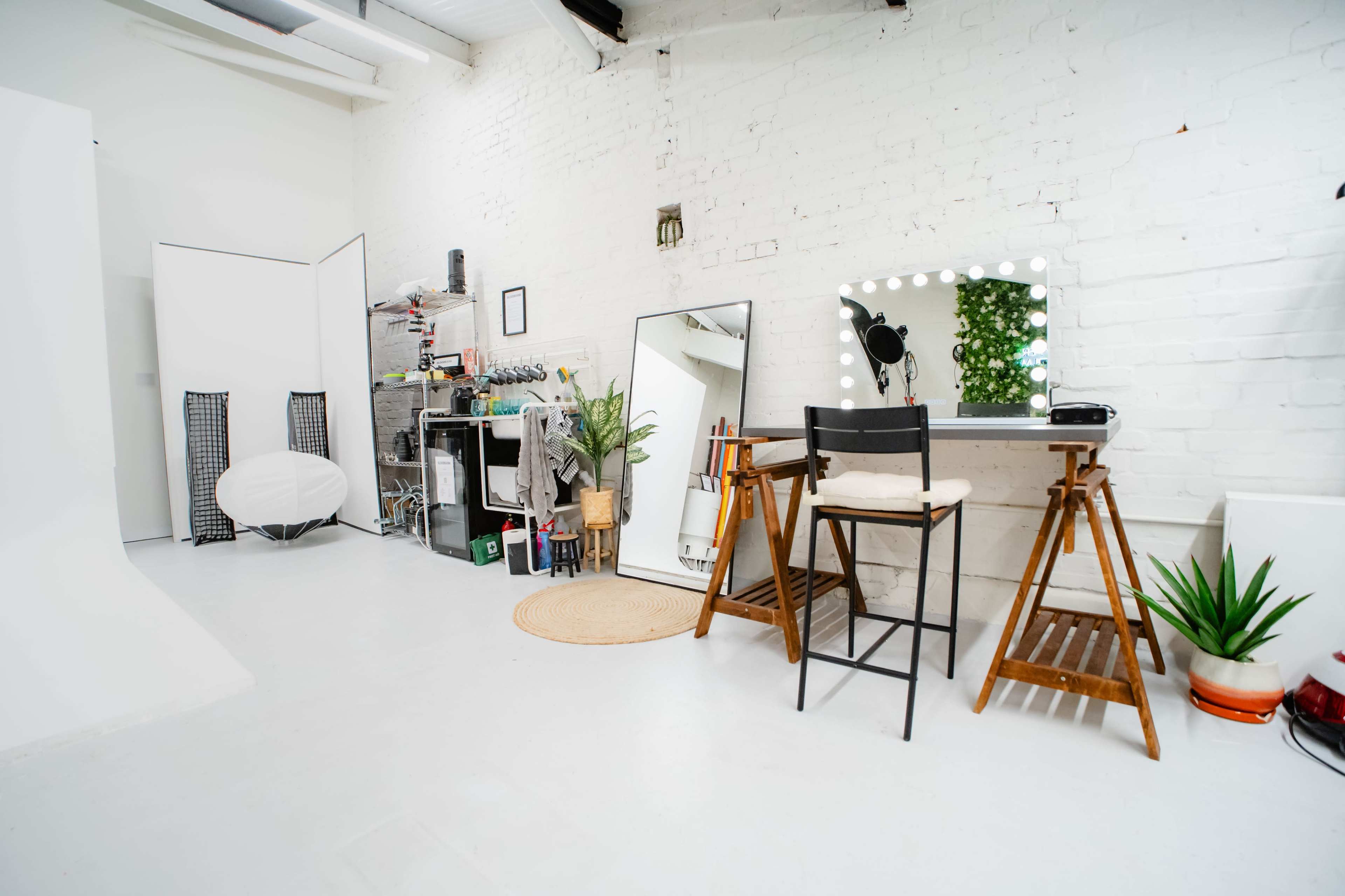 The image shows a bright studio space featuring a vanity mirror with lights, a wooden chair, a large mirror, and various props and plants arranged against white brick walls.