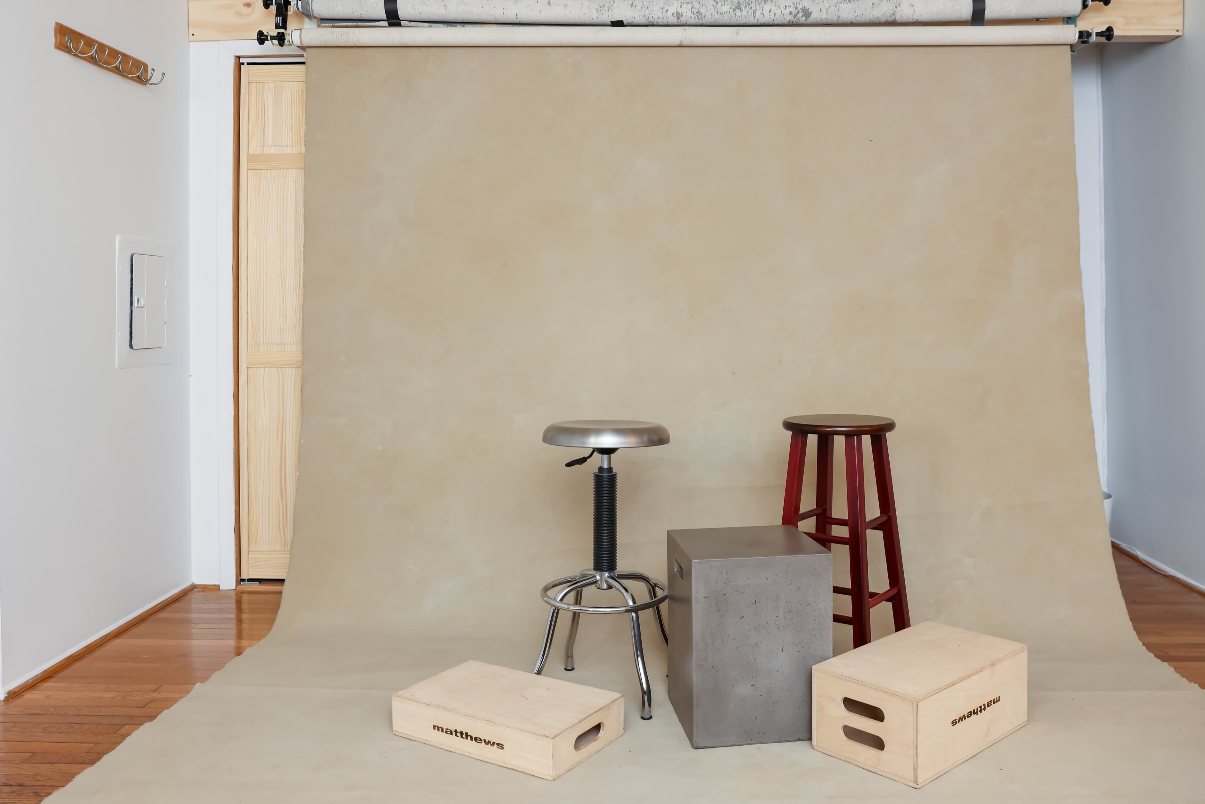 A minimalist studio setup featuring a beige backdrop with a metal stool, a wooden stool, a concrete block, and two wooden boxes arranged in front of it.